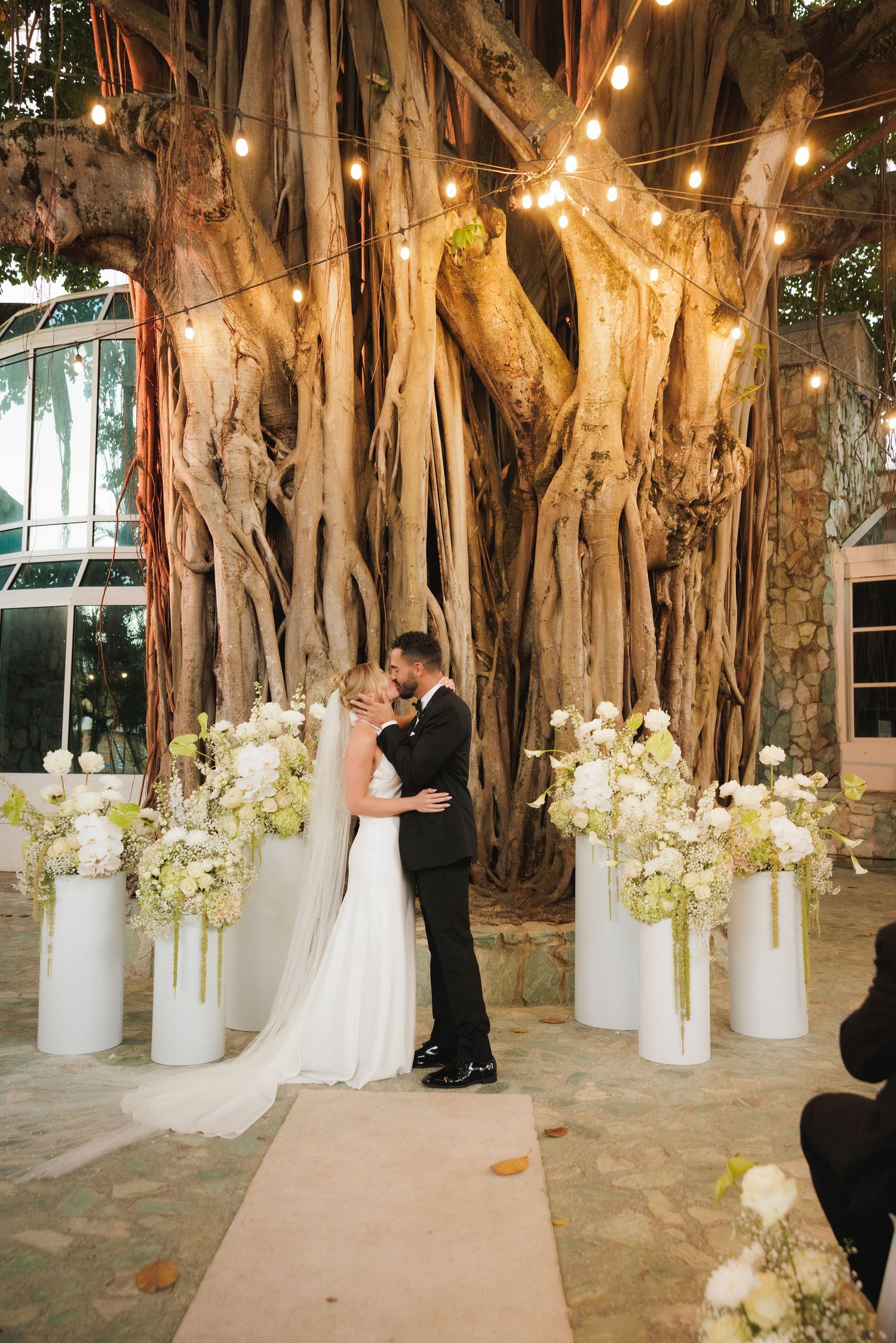 White floral arrangements line a romantic garden ceremony space beneath a majestic banyan tree illuminated by string lights.