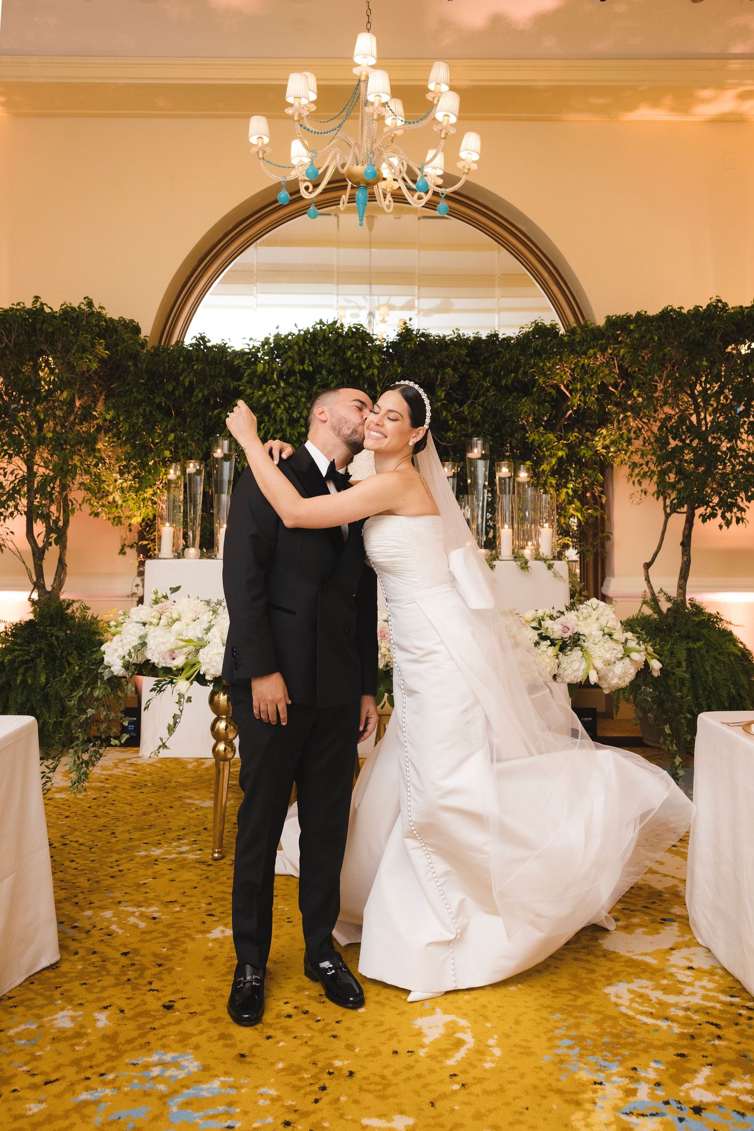Wedding couple dances and twirls in an elegant ballroom decorated with green trees and golden autumn leaves on the floor.