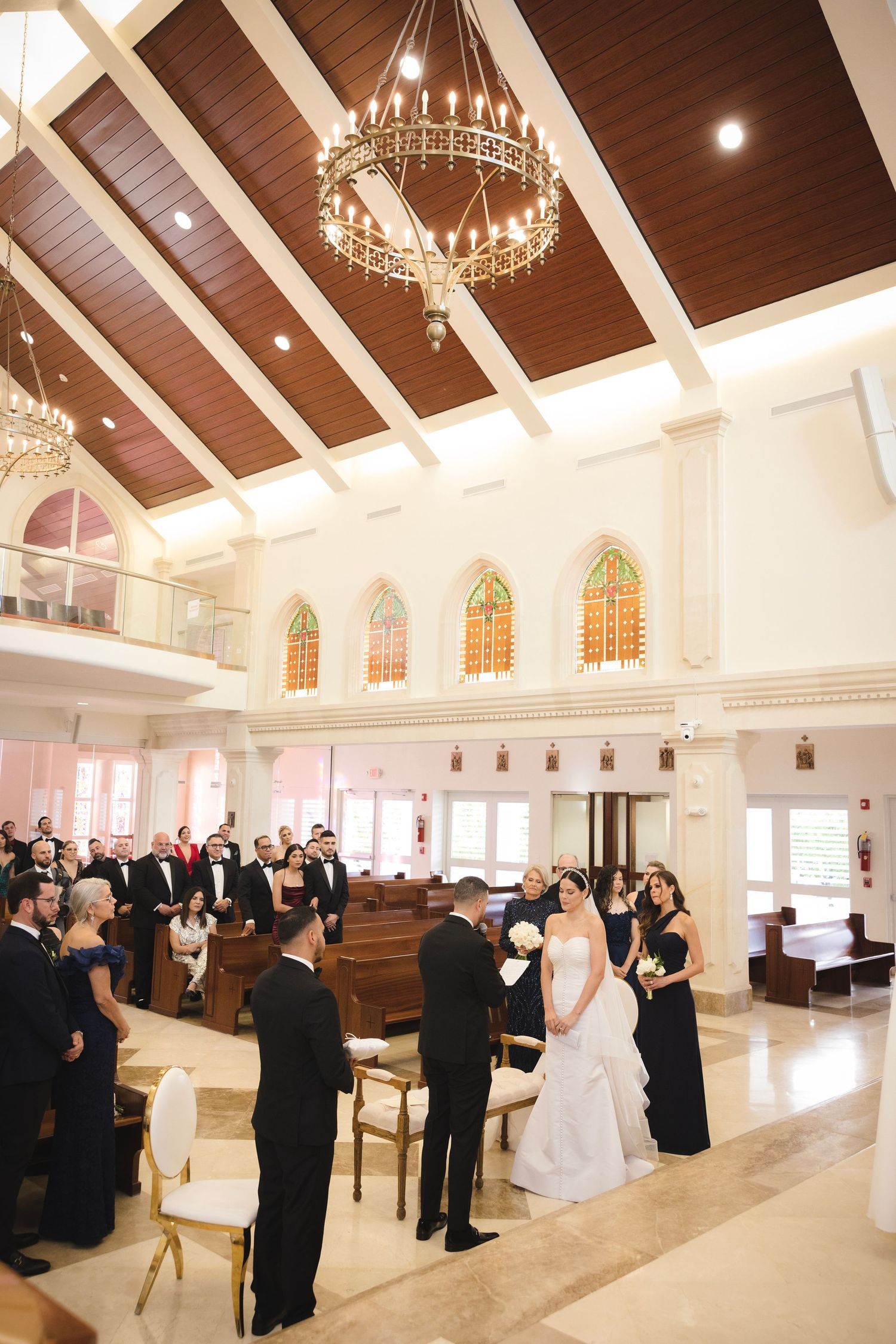 A grand church interior with stained glass windows and ornate chandelier hosts an elegant wedding ceremony.