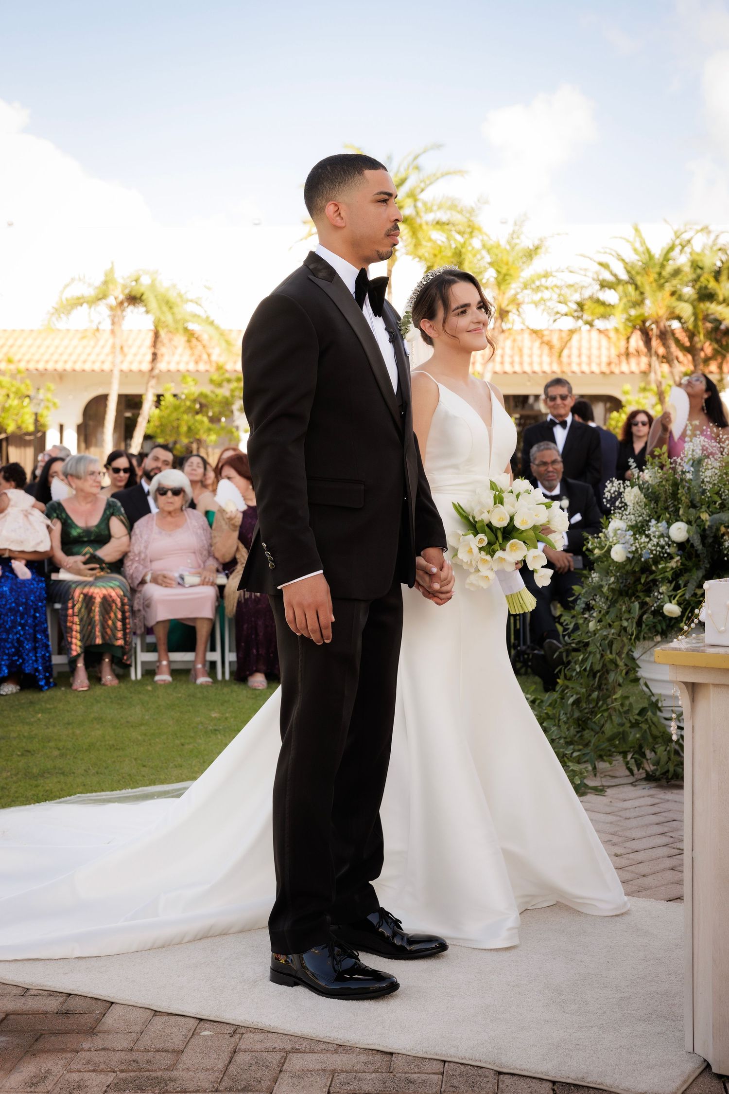 Wedding couple shares intimate moments during outdoor ceremony on stone patio surrounded by lush greenery and guests.