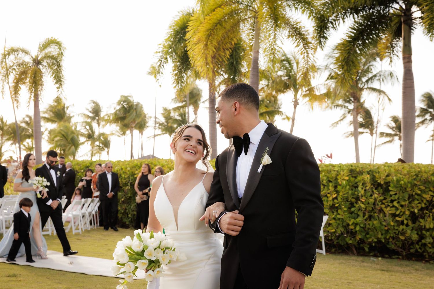 Couple shares romantic moments at sunset beach wedding with palm trees and ocean backdrop in elegant attire.