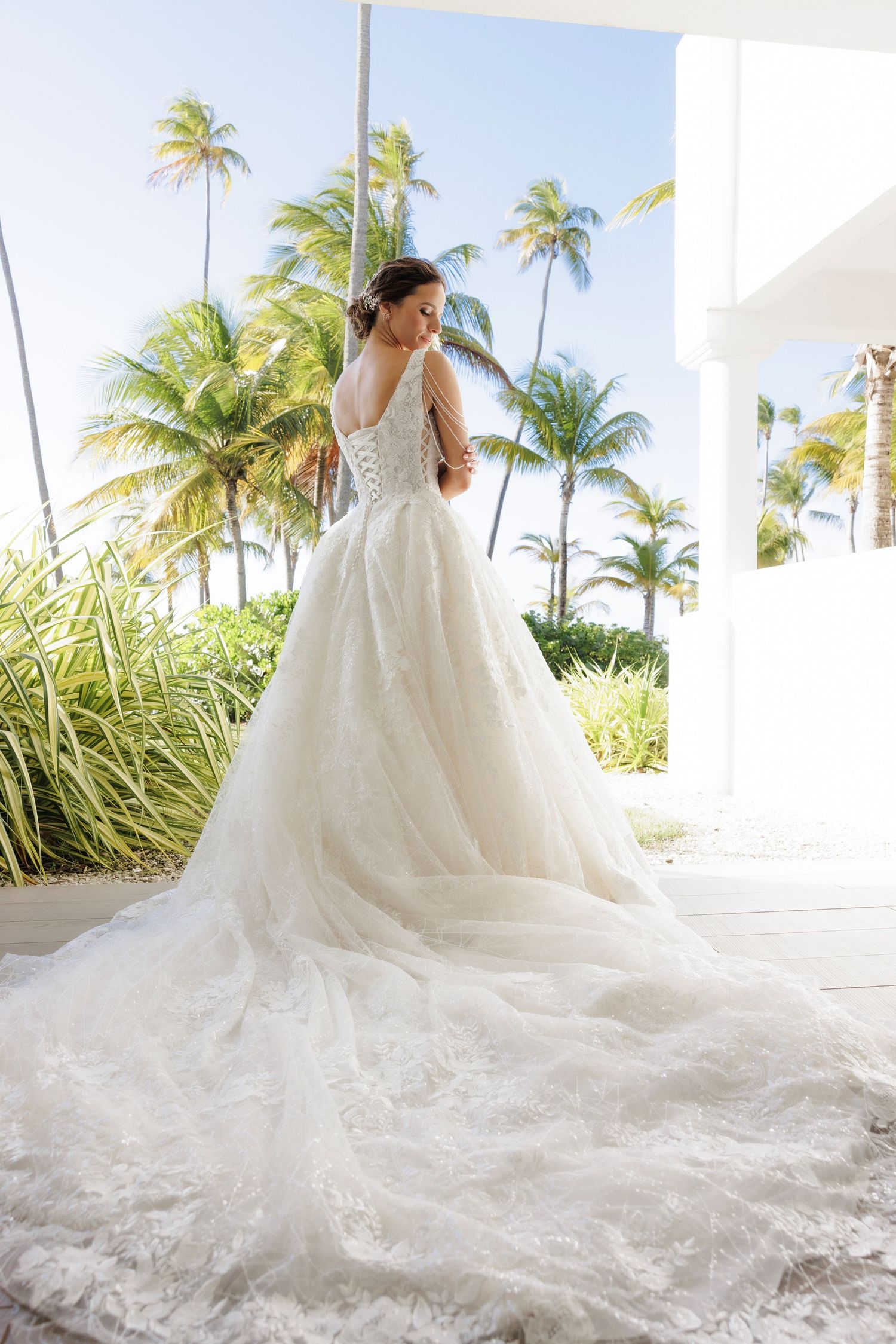 A stunning bridal gown with a long flowing train photographed against an elegant white corridor with palm trees outside.