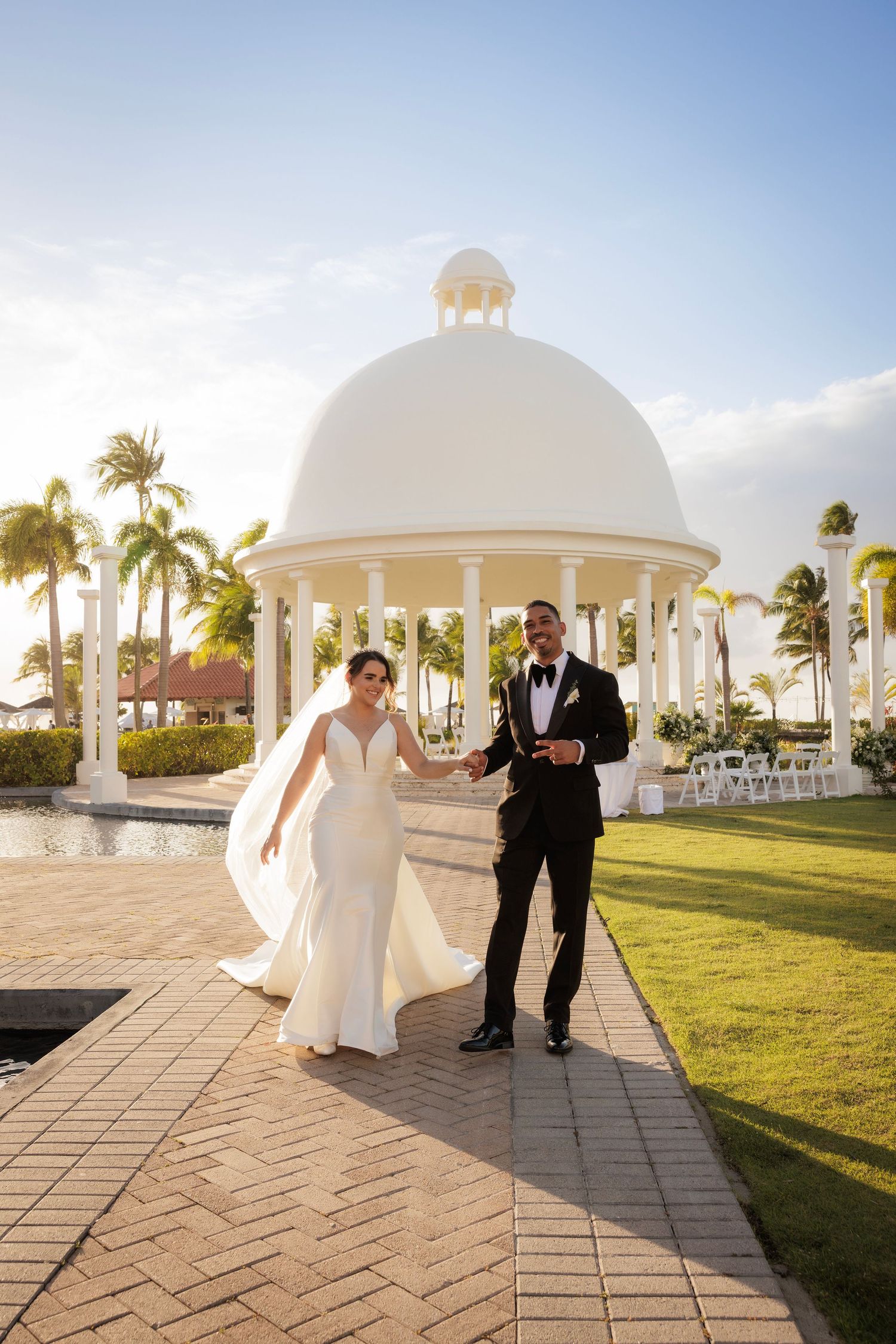 Elegant wedding couple walking at tropical resort with white domed gazebo and palm trees at sunset.