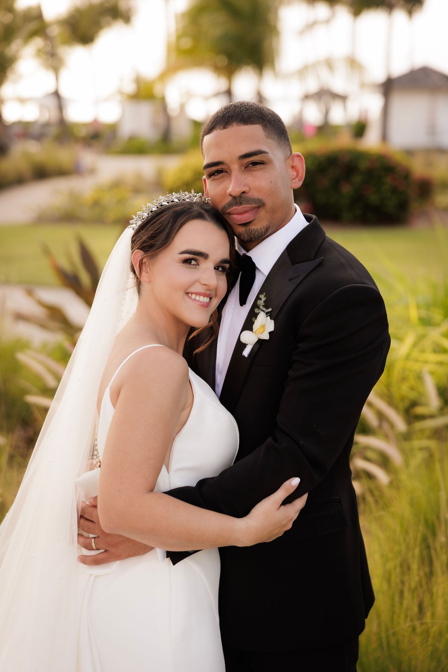 A scenic outdoor wedding portrait of a couple embracing in formal attire surrounded by lush greenery at sunset.