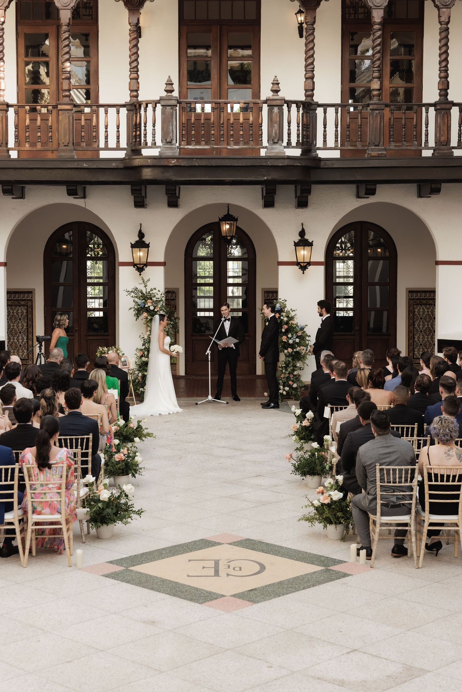 Wedding guests gather in an elegant Spanish colonial style courtyard with arched columns and balconies.