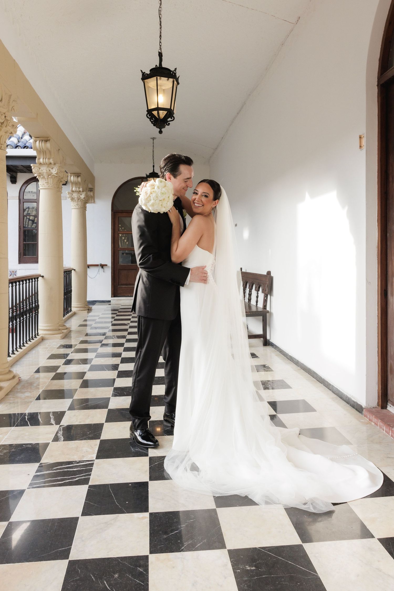 Newlyweds share intimate moments in a colonnade with dramatic checkerboard flooring.