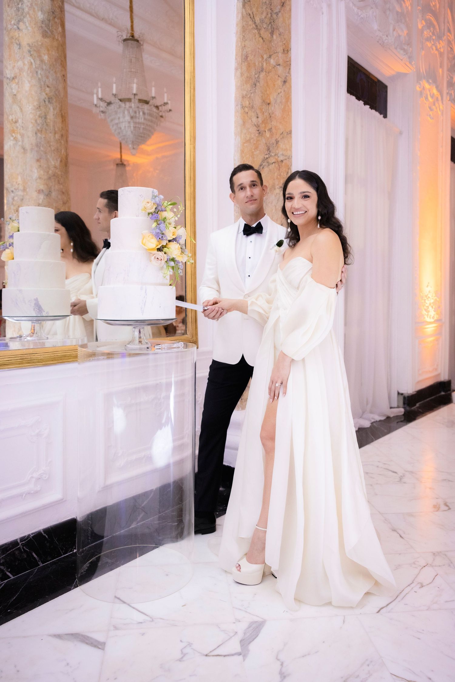 Wedding couple posing beside their elegant tiered white cake in a marble-columned venue.