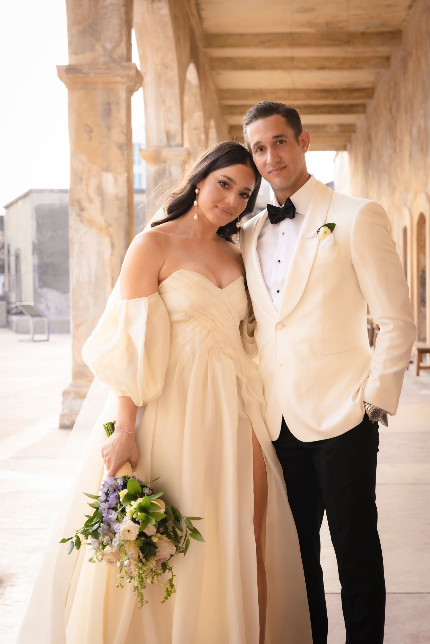 Wedding couple poses elegantly under stone colonnade wearing white dress and black tuxedo.