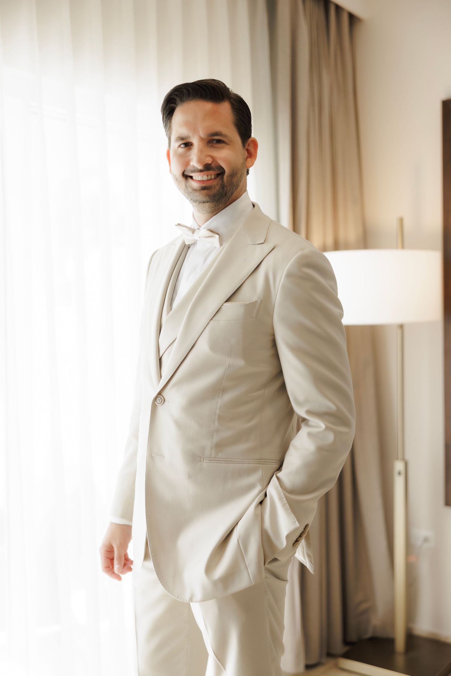 A groom wearing a white formal suit stands in front of curtains in a sunlit room with a cheerful demeanor.
