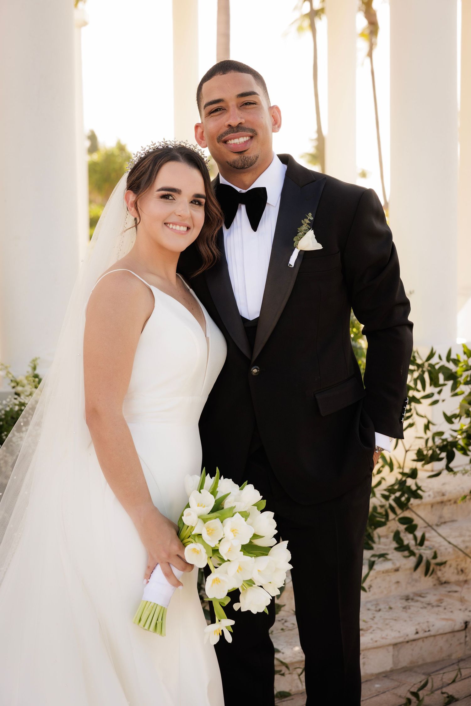 A happy bride and groom pose for wedding portraits in front of white columns while holding a bouquet of white flowers.