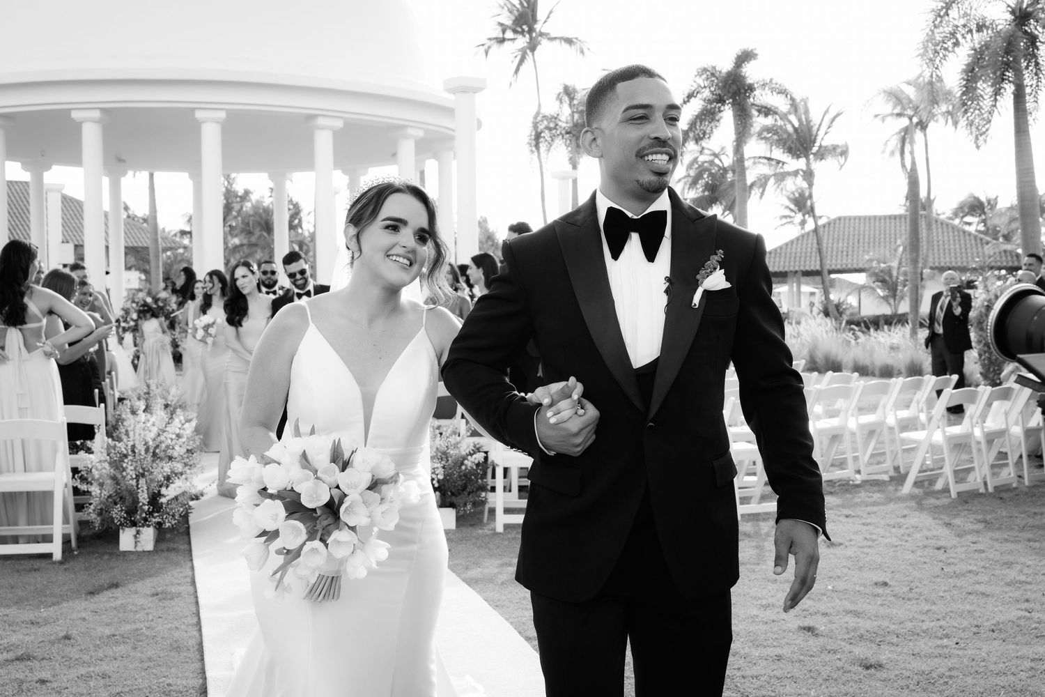Newlyweds and wedding party in formal attire walk together outdoors at a tropical resort during a sunset celebration.