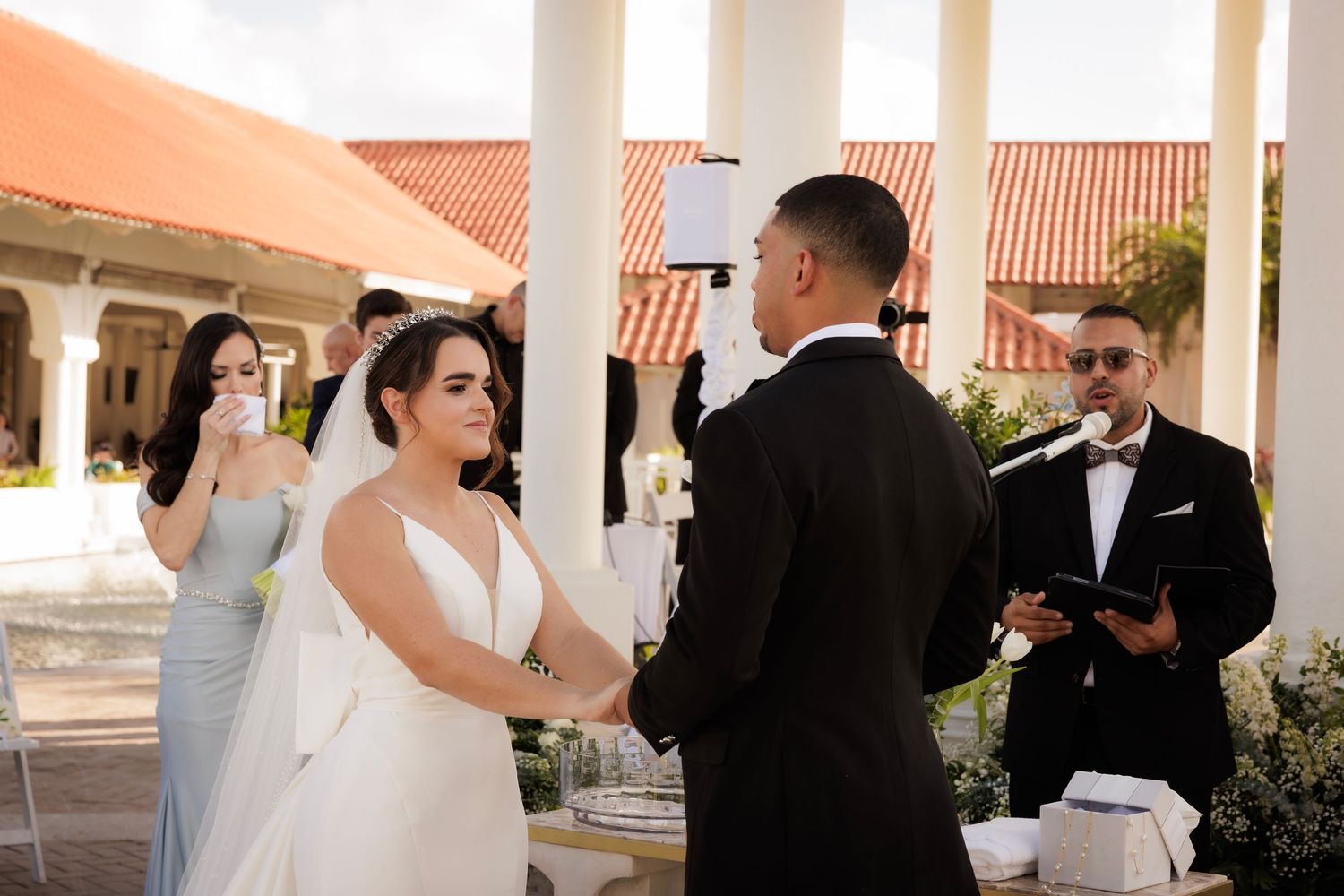 A wedding ceremony takes place under a covered outdoor terrace with terra cotta roofing in the background.