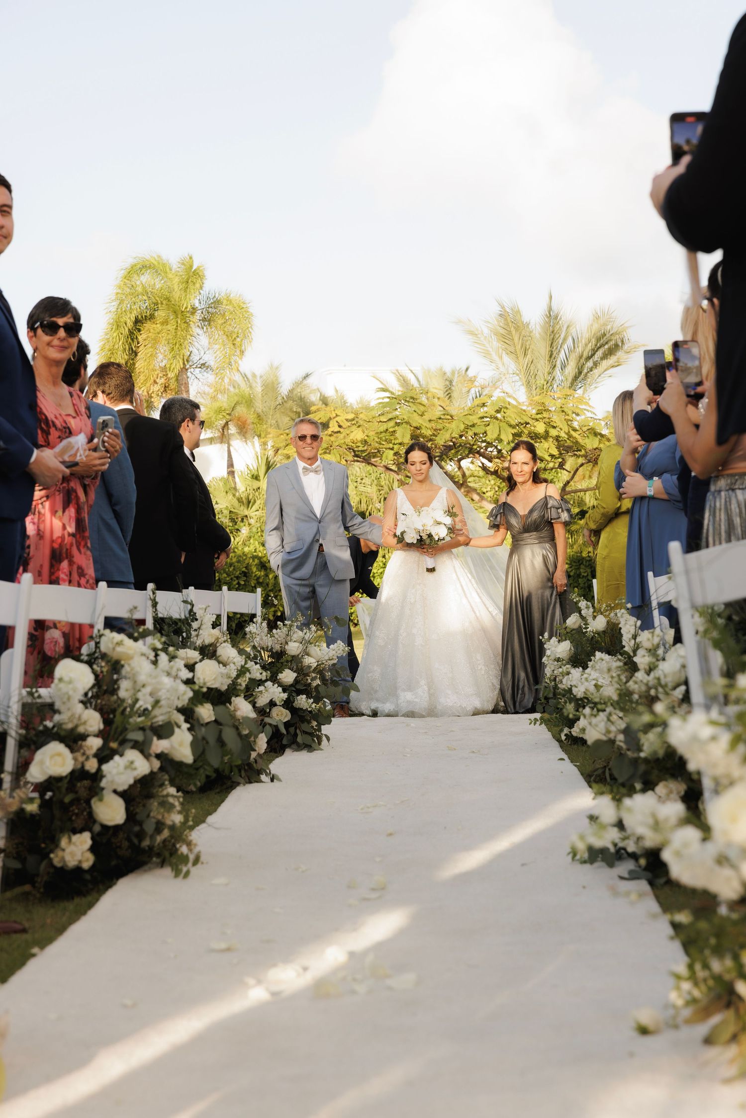 Beautiful white floral arrangements line a wedding aisle with palm trees and guests visible in the background.