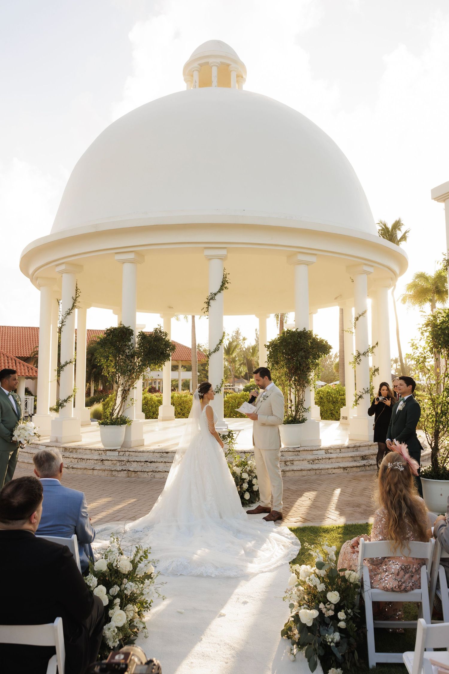 A series of white gazebos in an outdoor wedding venue with floral decorations and guests at sunset.