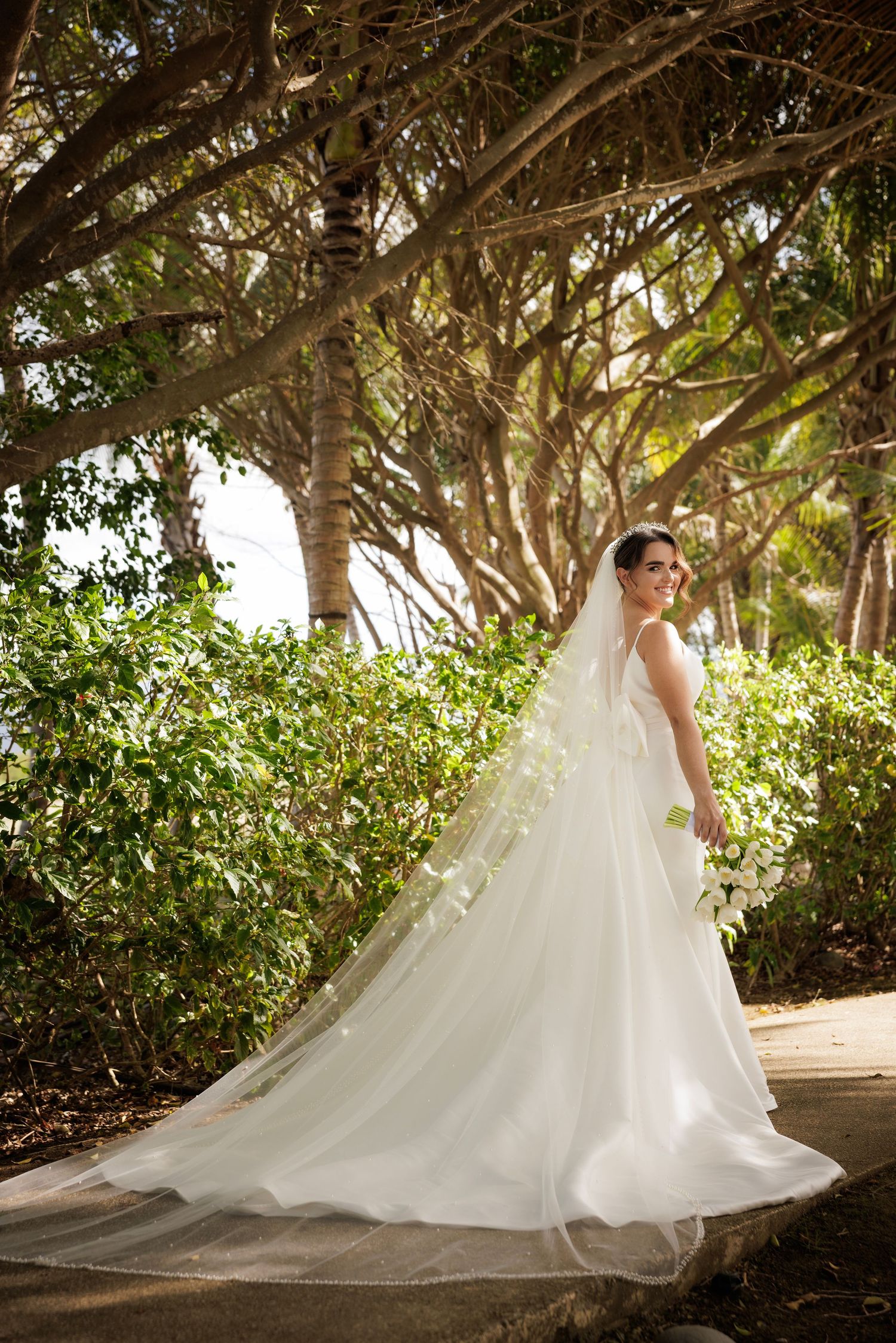 A bride in a flowing white wedding gown poses in a garden path underneath arching trees with dappled sunlight.