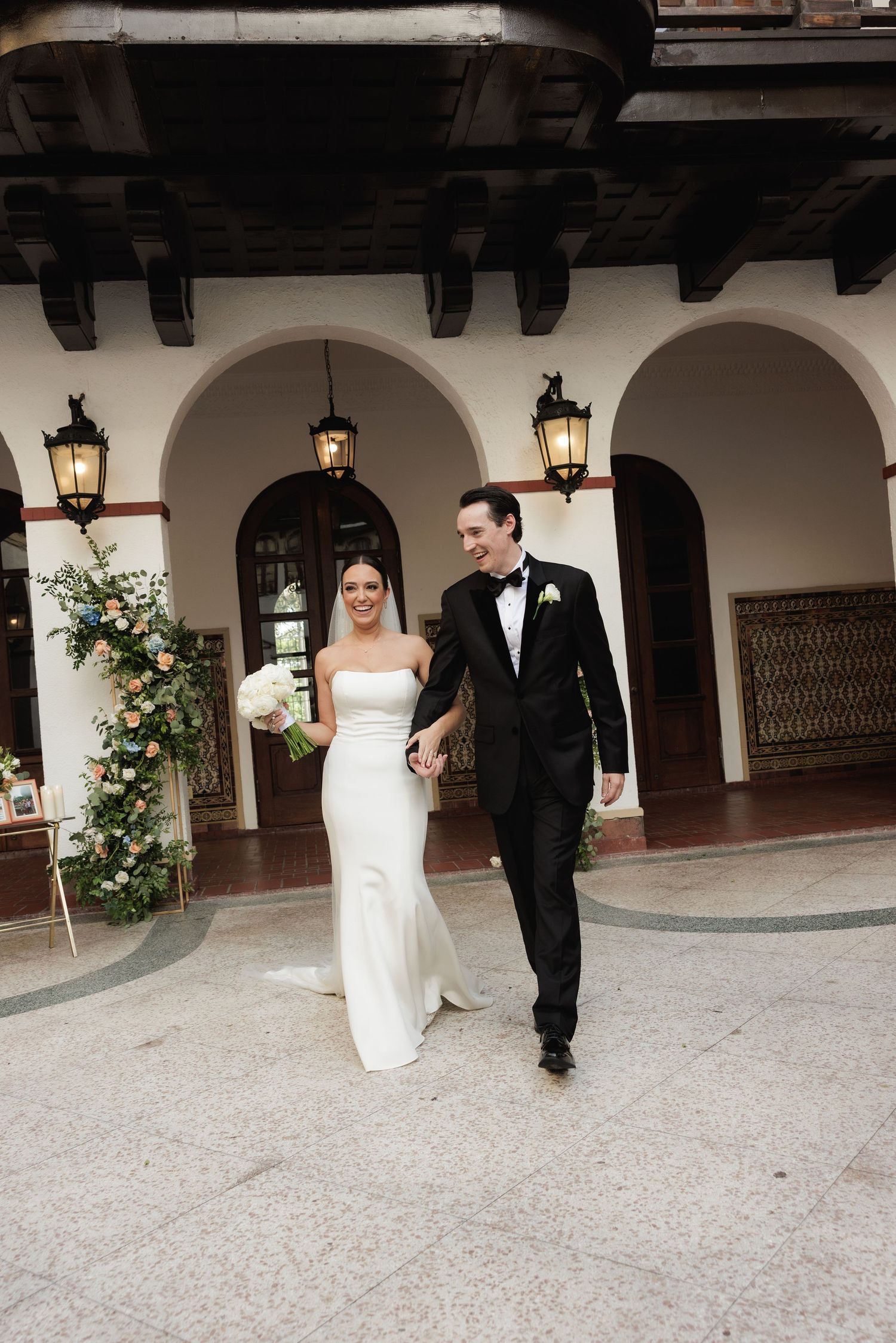 A bride and groom walk together through an elegant Spanish-style colonnade decorated with floral arrangements and lanterns.