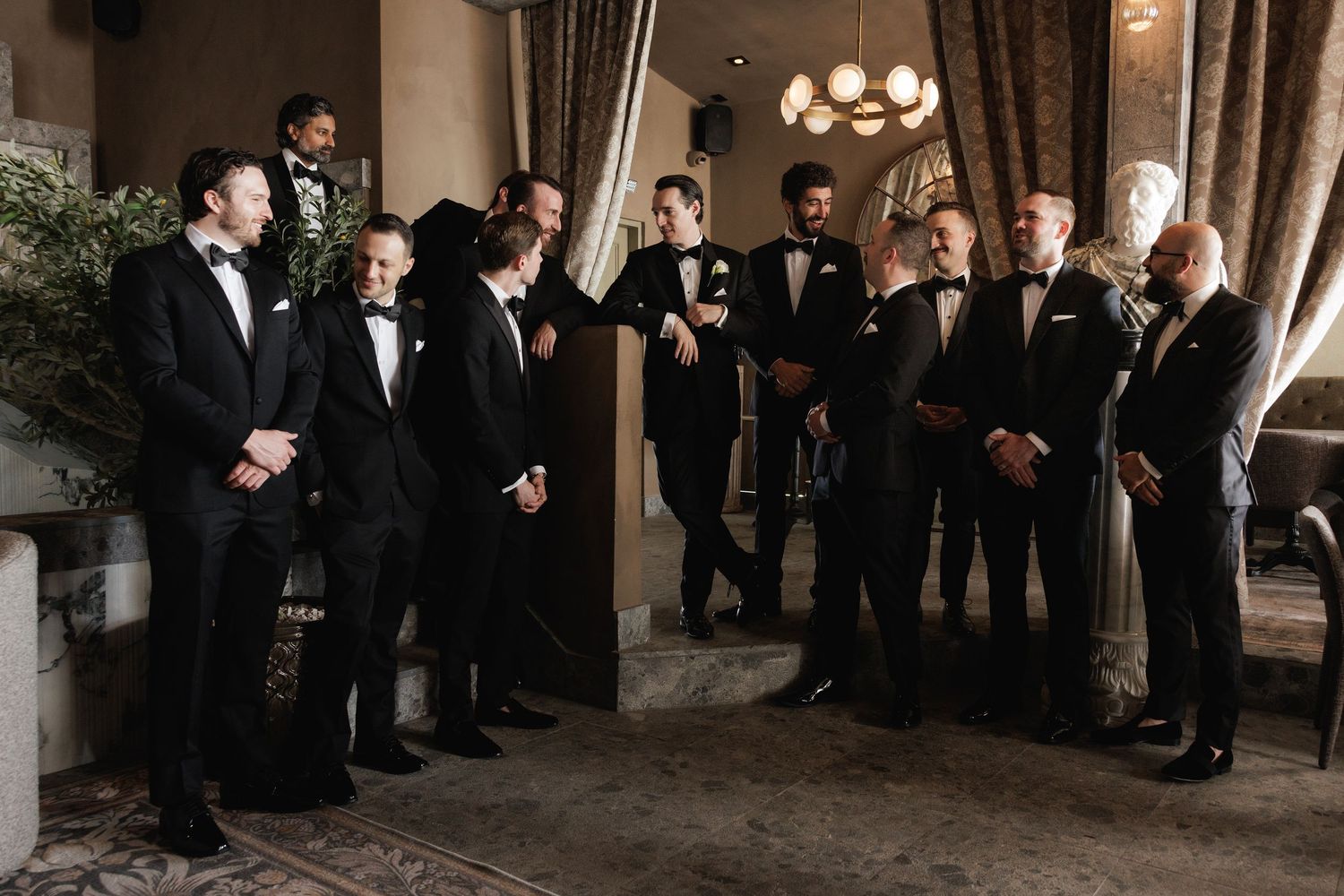 Groomsmen in black tuxedos gather for wedding party photos in an elegant indoor venue.