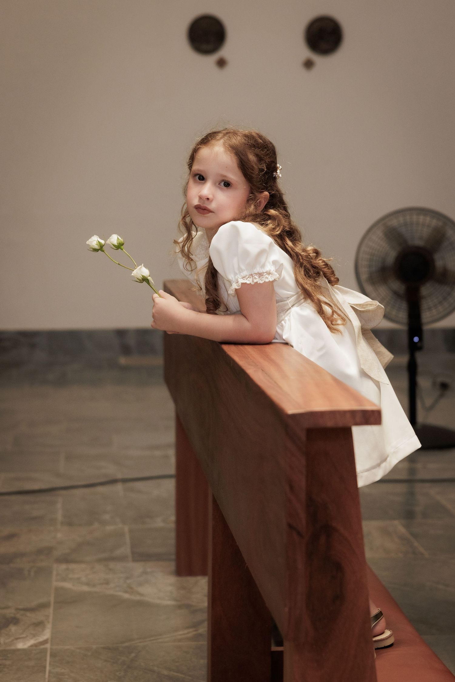 A young flower girl in a white dress peeks over a wooden church pew.