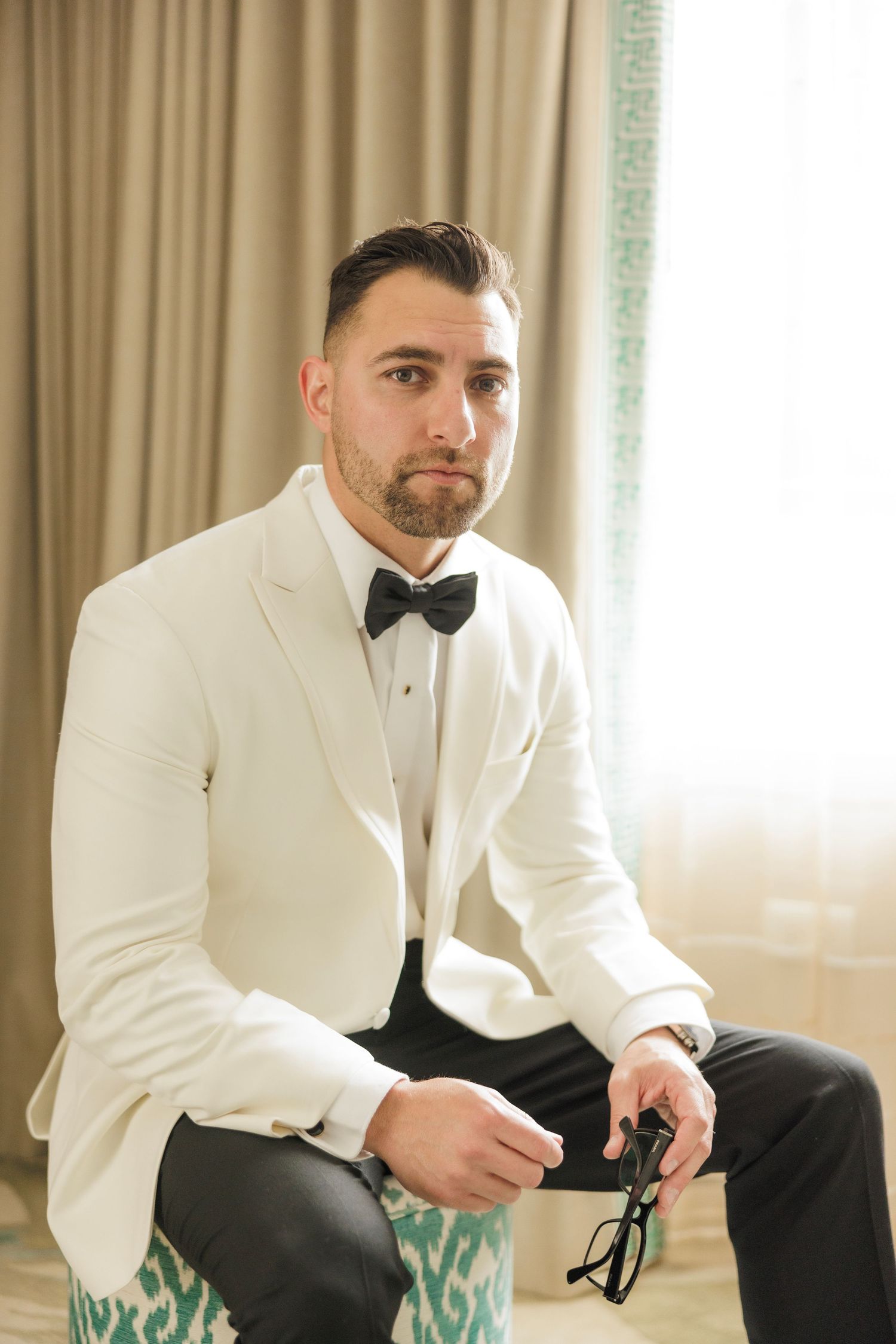 Groom seated in white dinner jacket and bow tie holding sunglasses in hotel room.