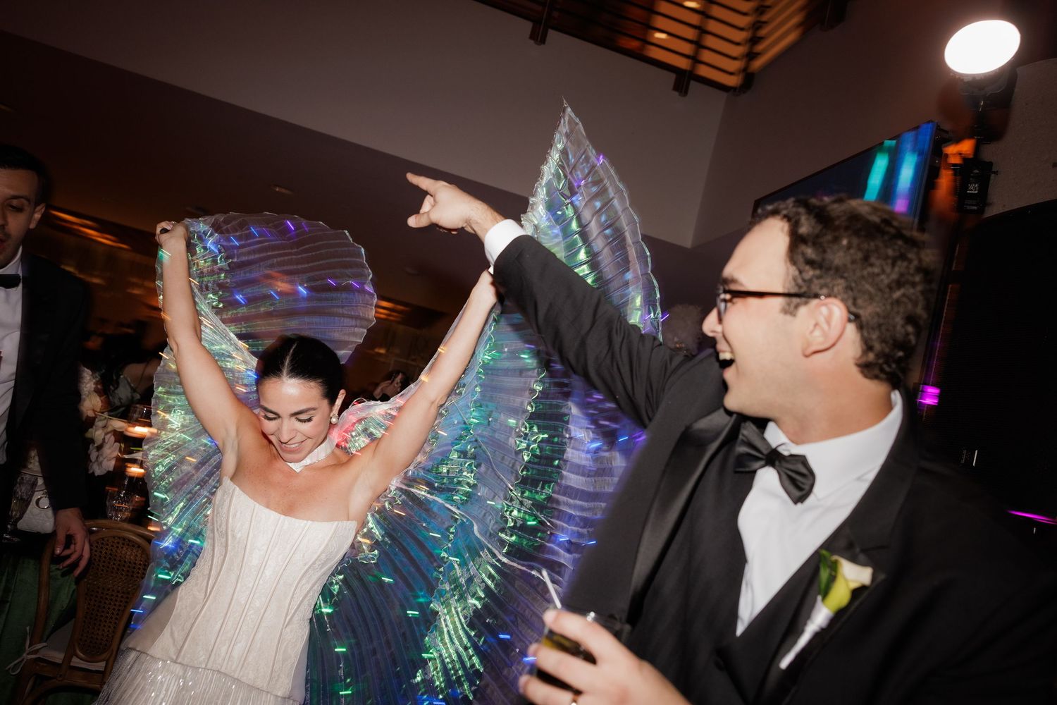 Couple dancing energetically at a wedding reception with shimmering LED butterfly wings creating magical lighting effects.