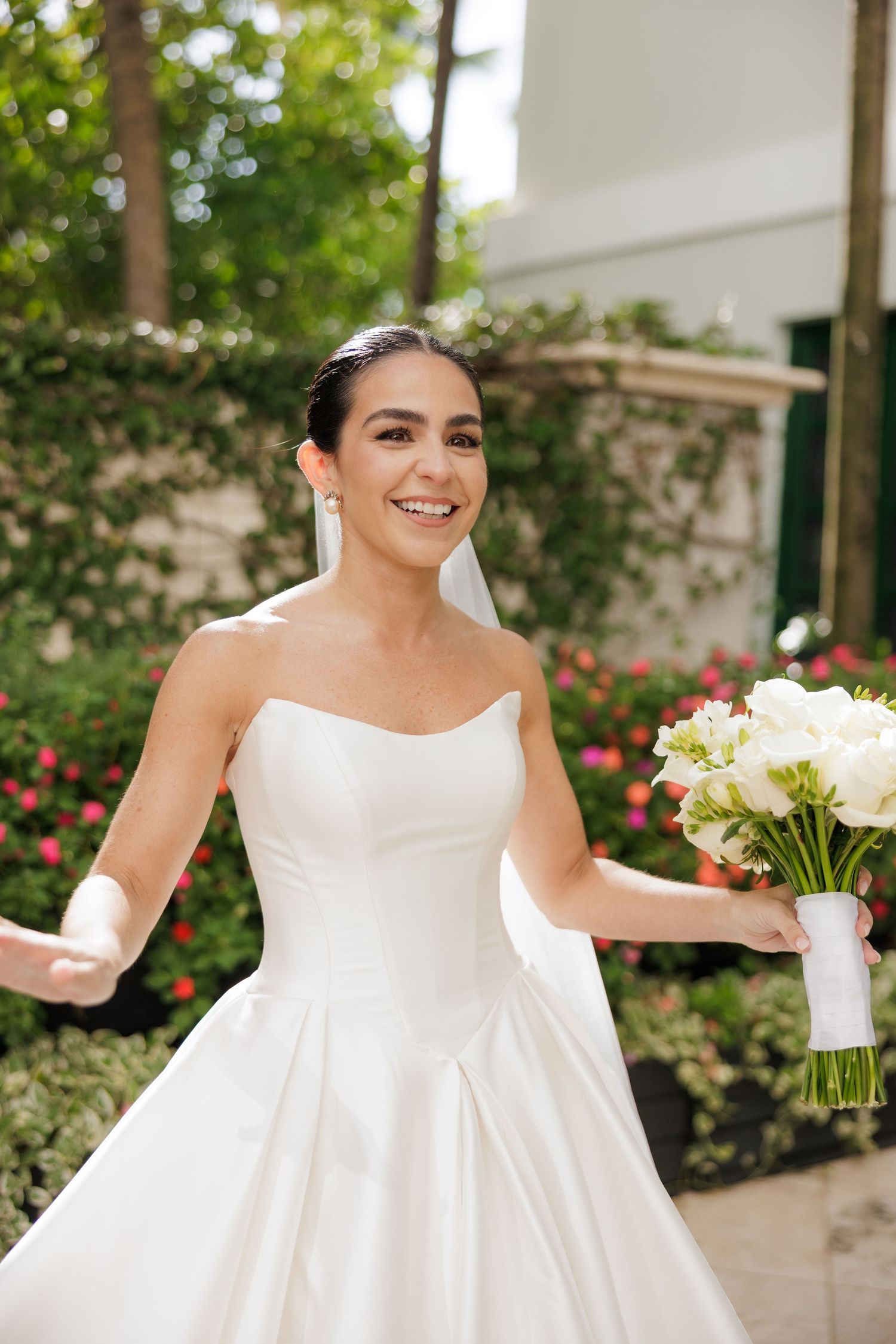A bride in a classic white strapless wedding gown smiles while holding a white floral bouquet in an outdoor garden setting.