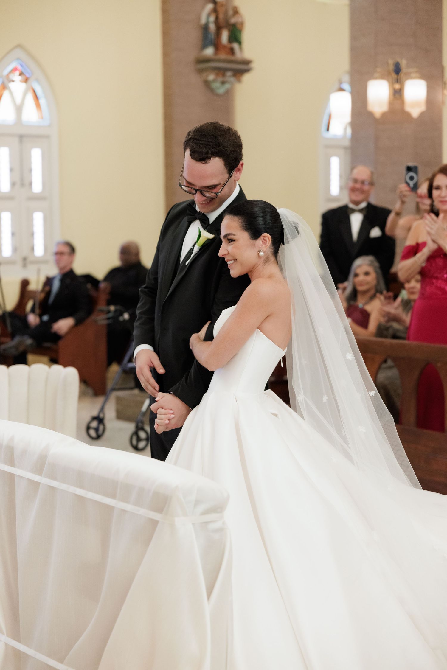 Bride in flowing white gown walks through church aisle with veil trailing behind.
