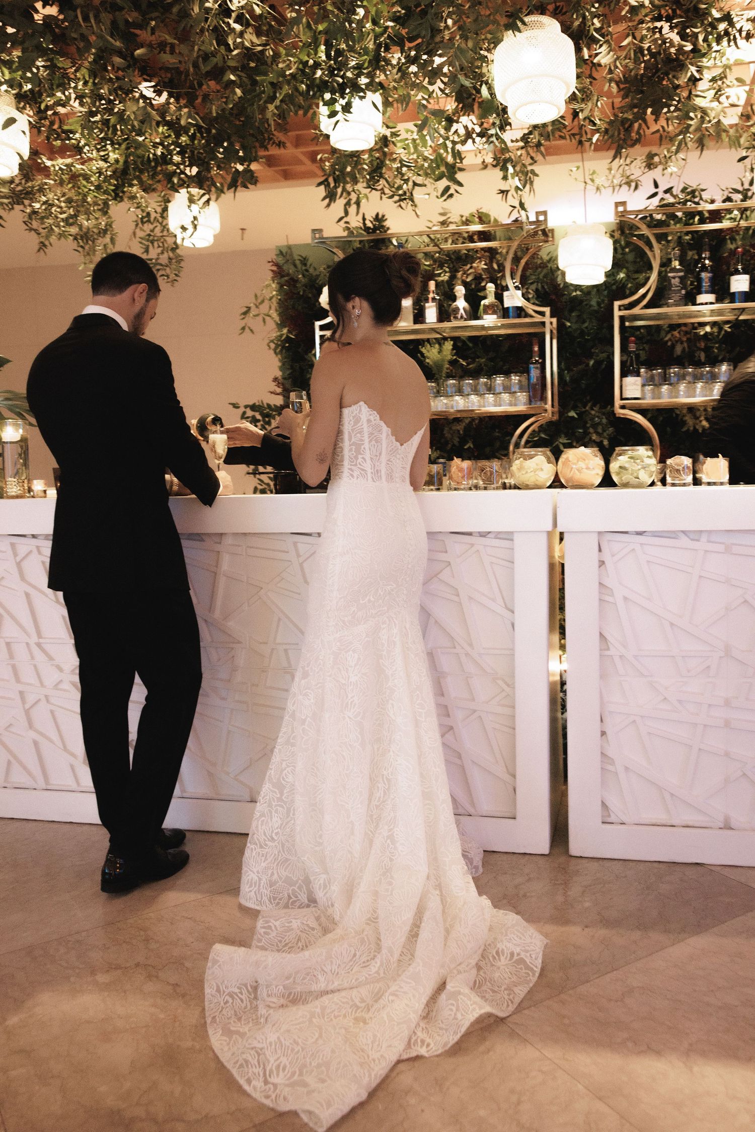 A bride in a flowing lace gown stands with her companion at an elegant marble bar decorated with hanging lanterns and greenery.