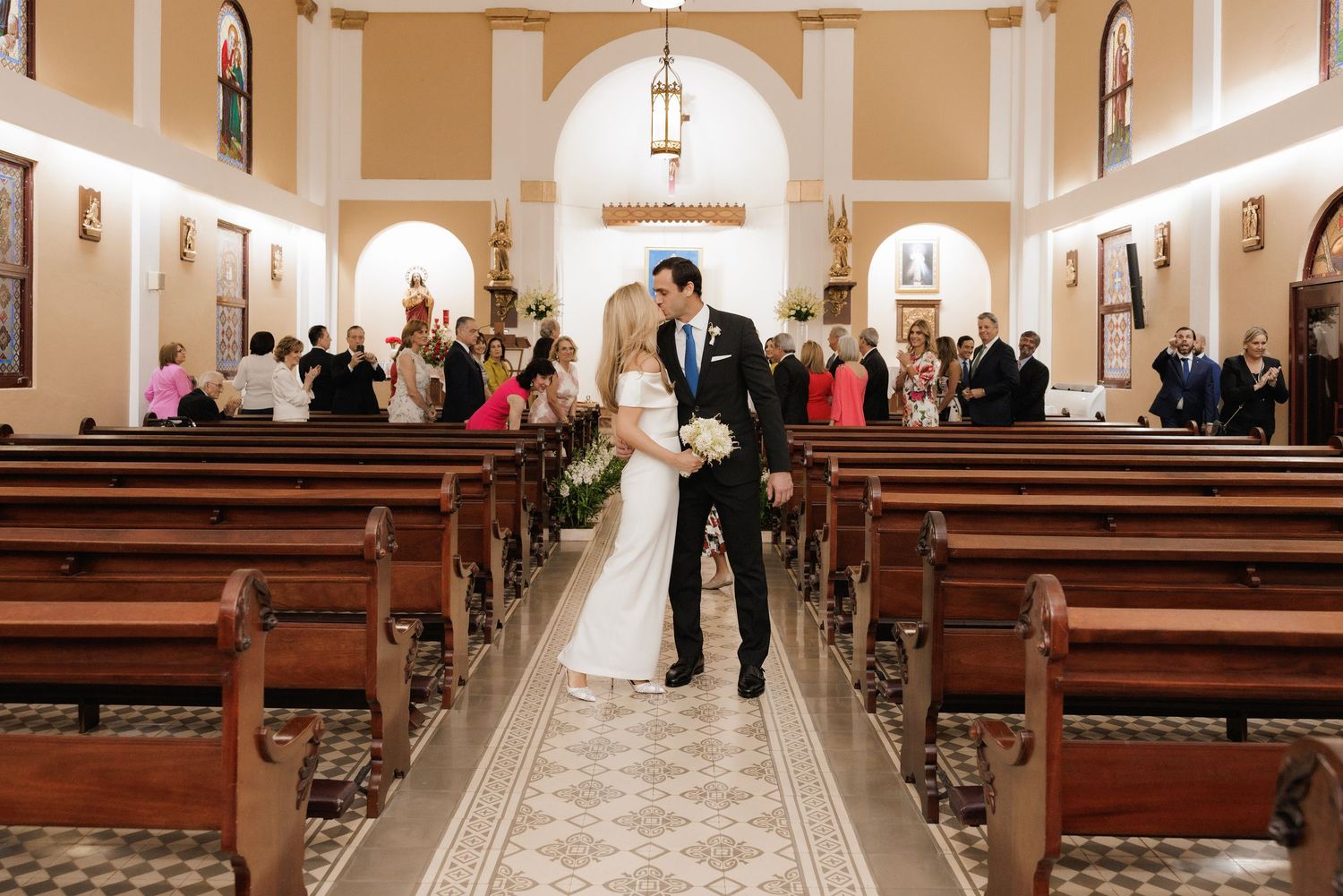 A wedding processional takes place inside a grand church with tan walls and ornate lighting fixtures lining the aisle.