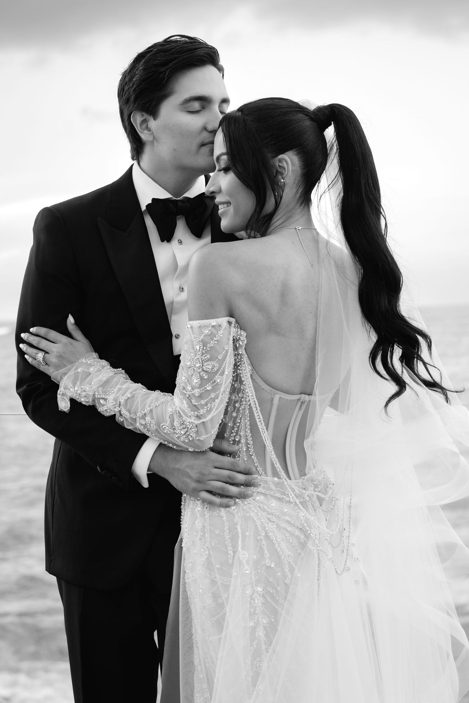 A romantic black and white wedding photo series showing tender moments between a couple in formal attire by the ocean.