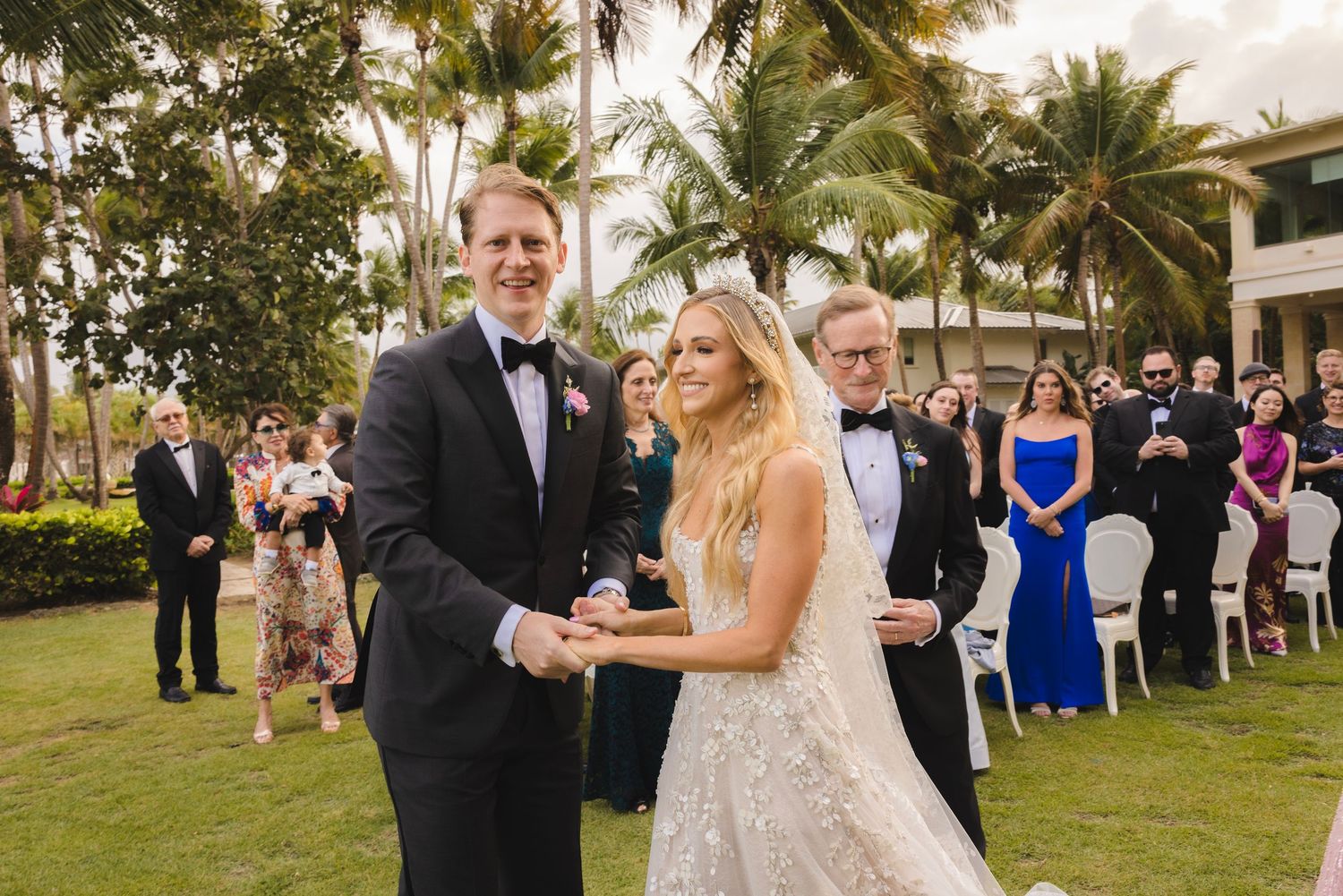 Two people share a joyful moment during an outdoor wedding ceremony on a palm tree-lined lawn with guests looking on.