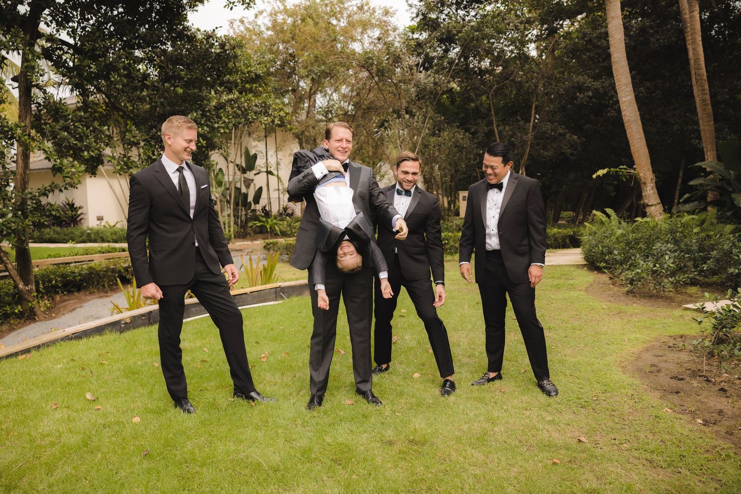 Groomsmen in black tuxedos pose playfully on a garden lawn during wedding day celebration.