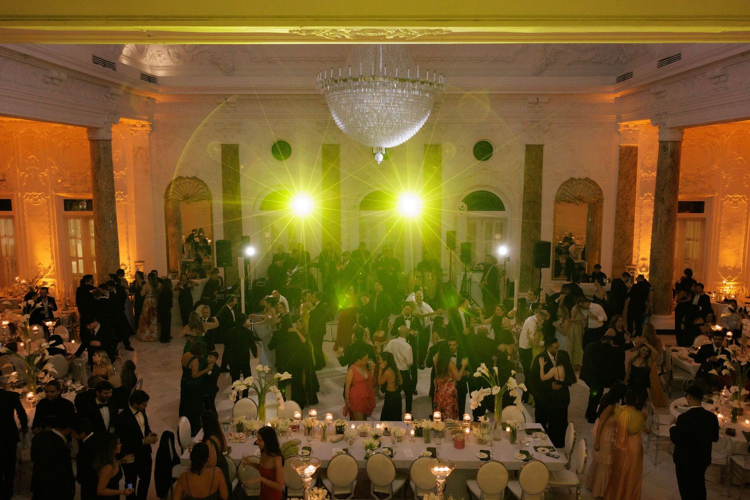 Wedding reception venue decorated with warm lighting and crystal chandeliers showing guests mingling on the dance floor.