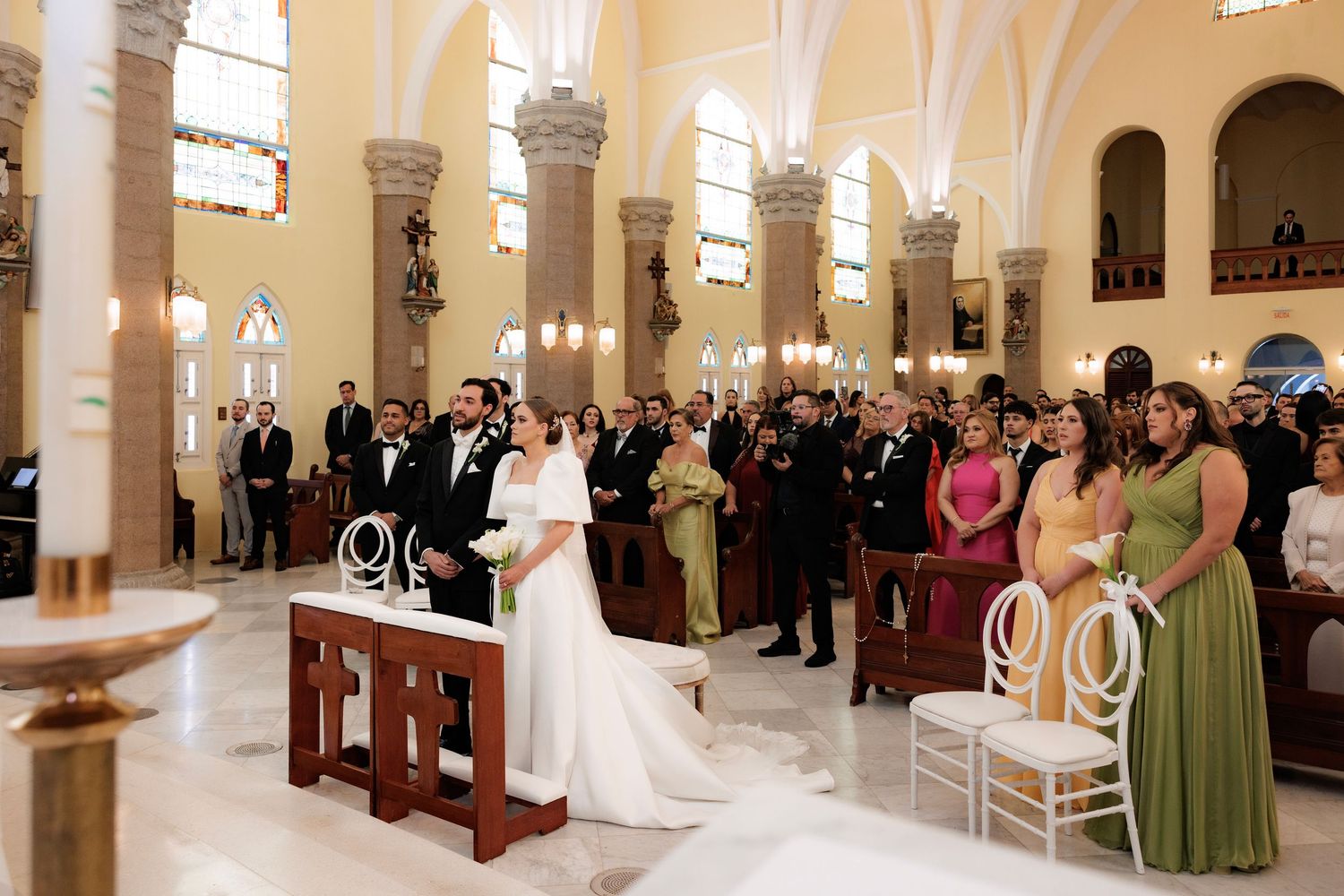 A traditional church wedding ceremony with guests gathered in a bright white interior with stained glass windows and arched columns.