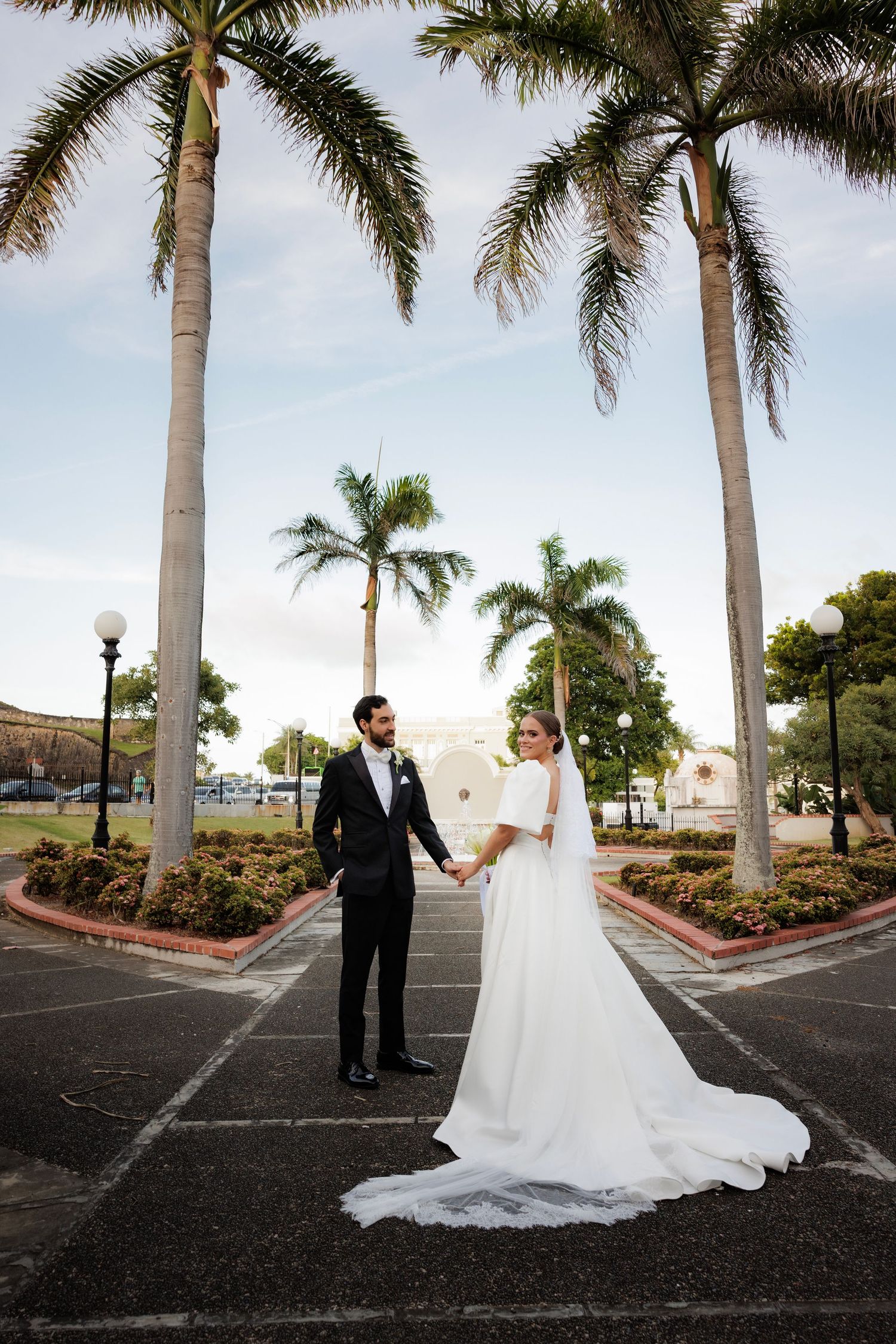 A bride and groom hold hands while standing in a tropical setting with tall palm trees and a cloudy sky.