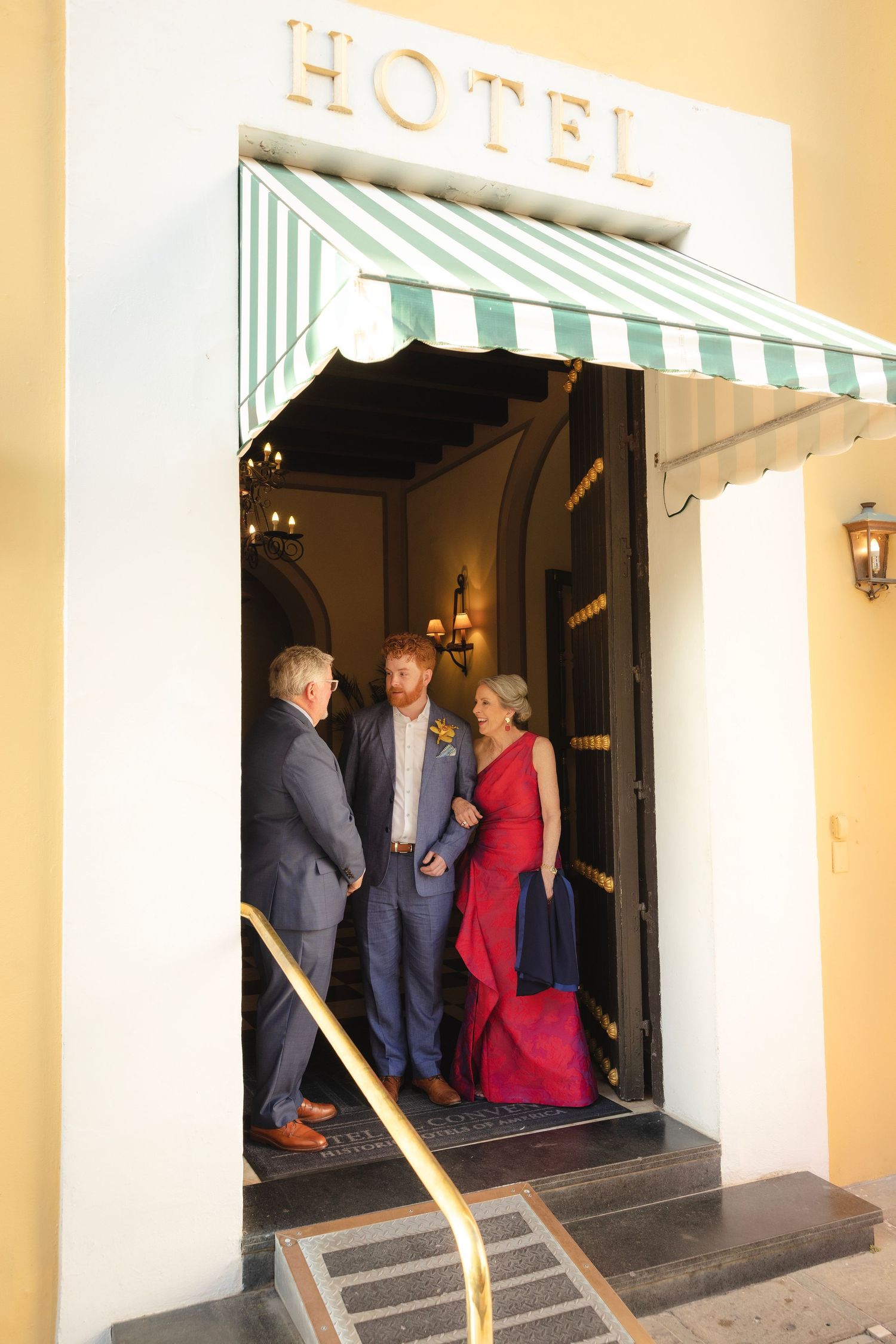 People in formal attire stand at the entrance of a hotel under a striped awning at dusk.