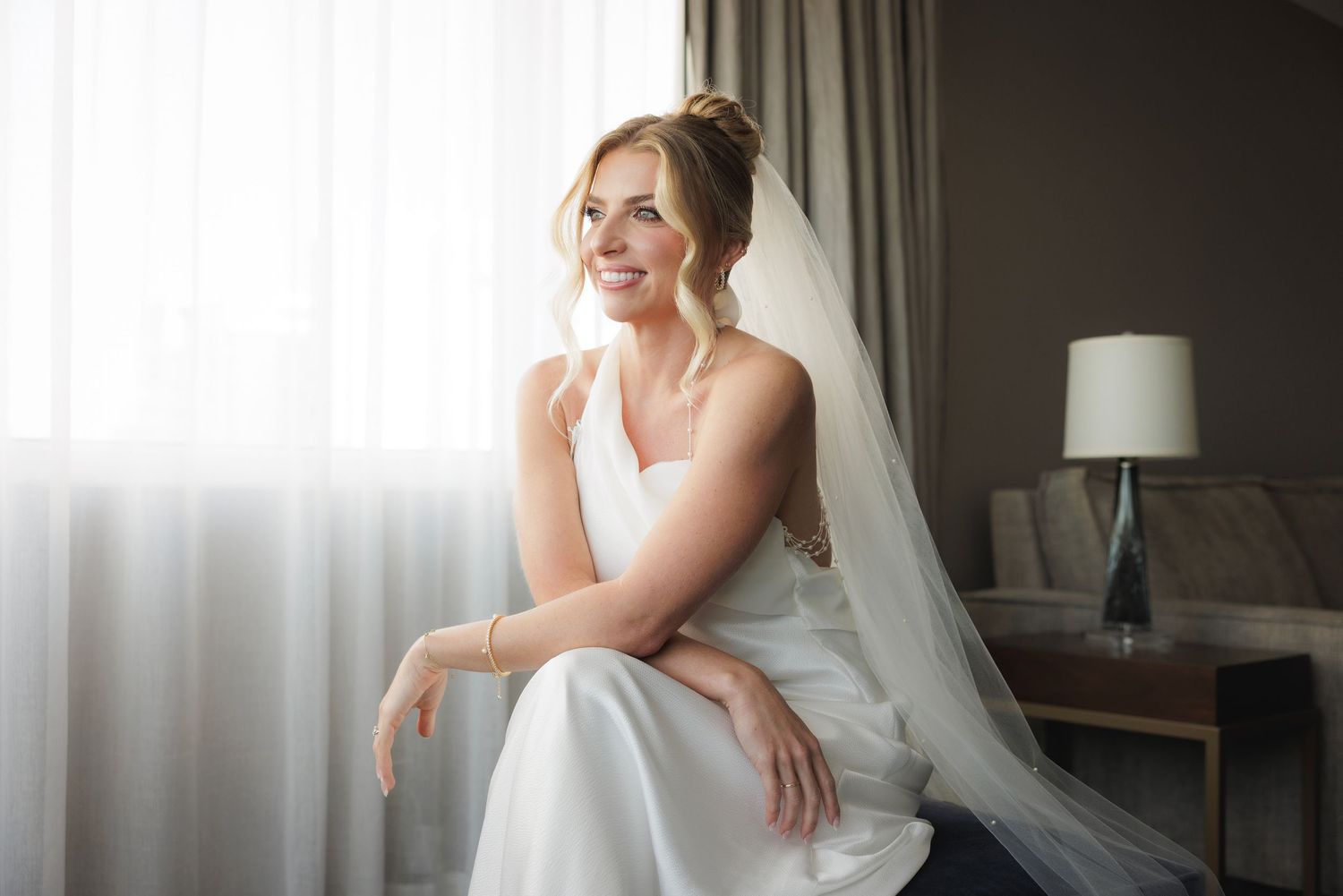 A bride in a white wedding dress sits elegantly on a couch in a hotel suite with natural window lighting.