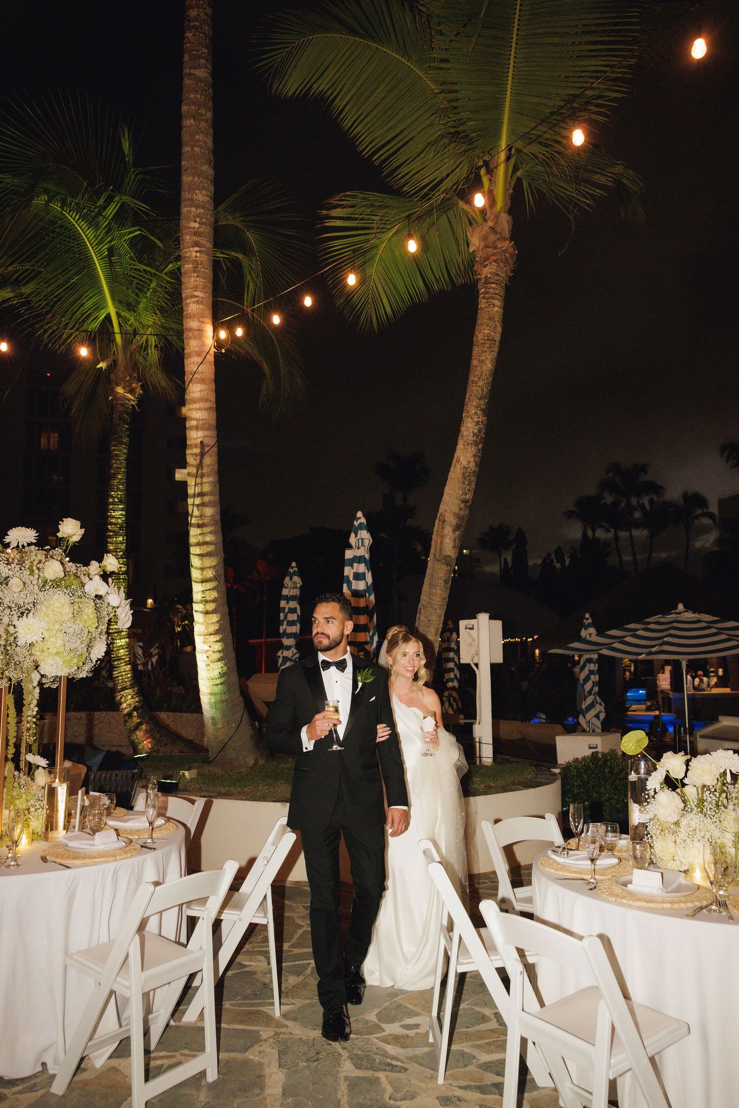 Outdoor evening reception setup with string lights, palm trees and elegant white table settings on a tropical patio.
