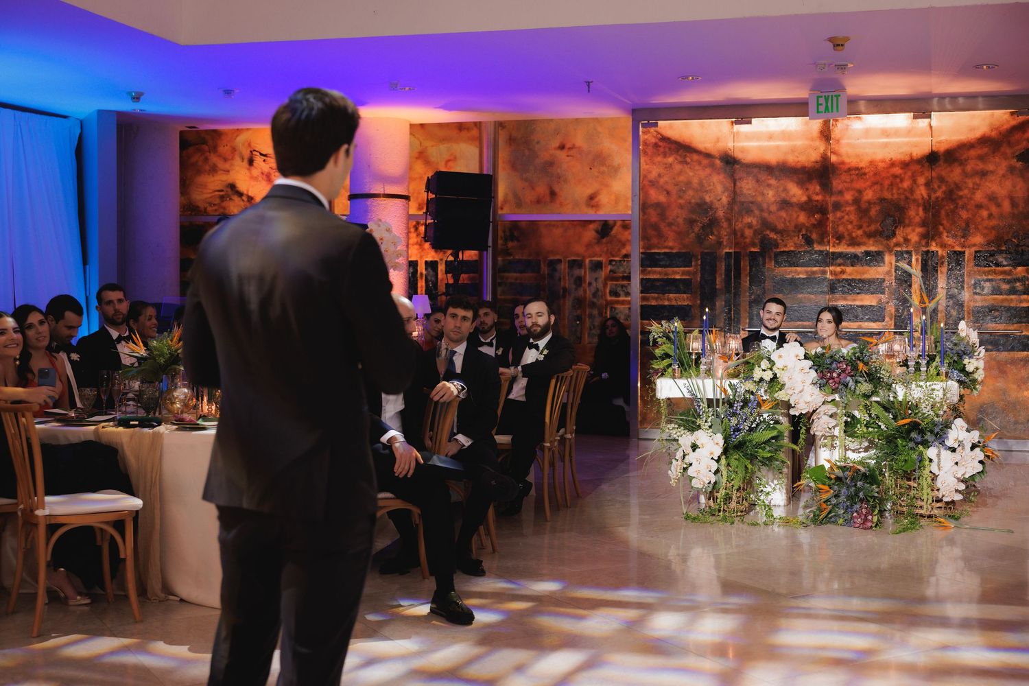 Guests mingle and dance at an elegant wedding reception with purple lighting and floral centerpiece decorations.