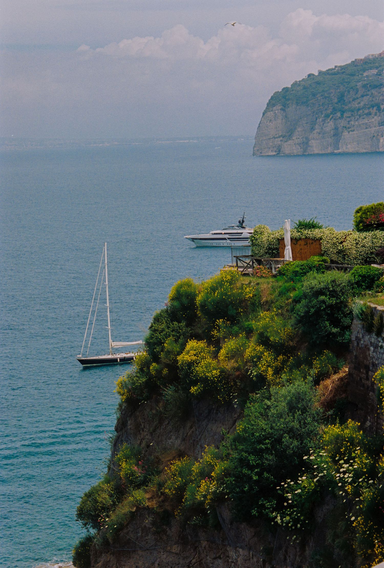 Scenic coastal cliffside with lush greenery, sailboat and yacht moored in calm blue Mediterranean waters near a rocky promontory.