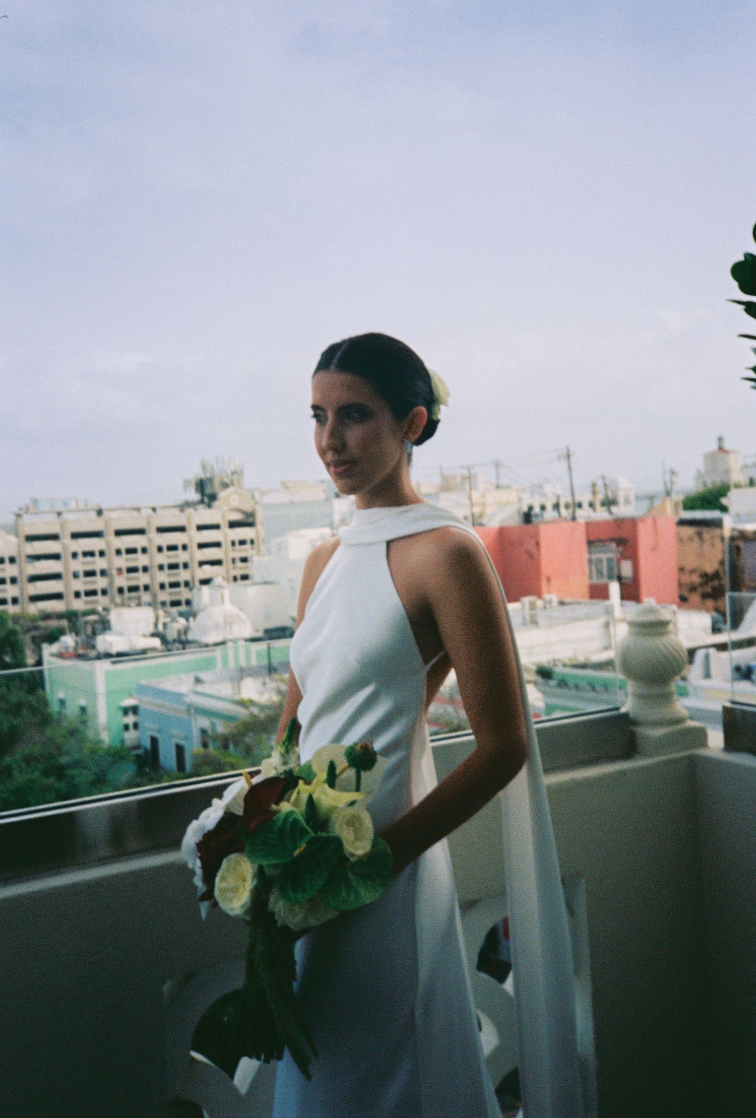 Elegant bride in white halter gown holding a green bouquet on a rooftop balcony with city views.