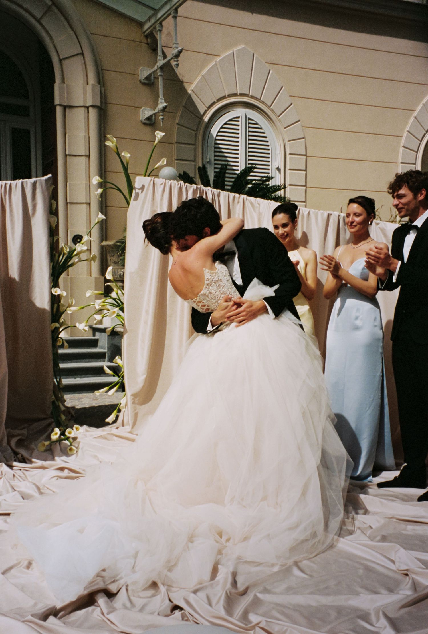 Bride and groom share first kiss at outdoor wedding ceremony with bridesmaids applauding.