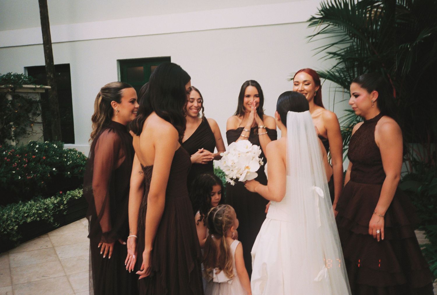 Bride in white gown holding bouquet surrounded by bridesmaids in dark burgundy dresses at an outdoor wedding venue.