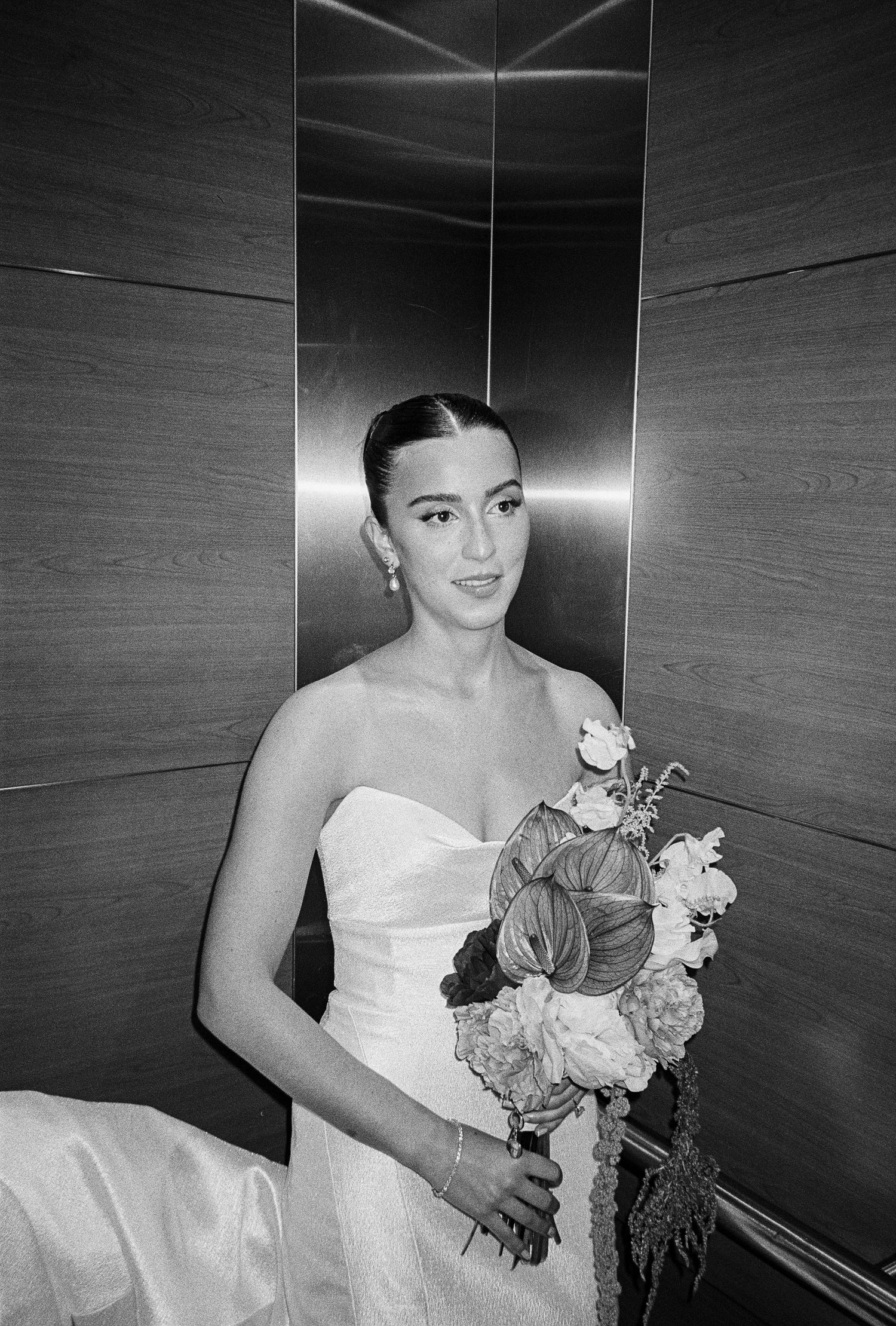 Elegant bride in strapless white gown holding a floral bouquet inside a modern elevator, black and white photo.