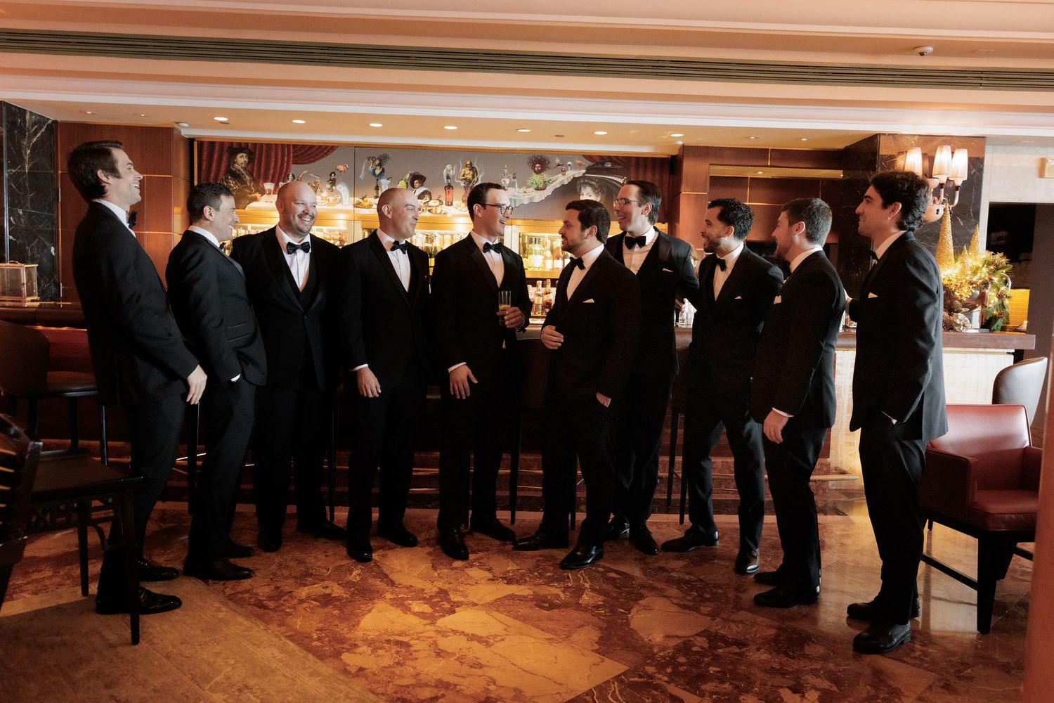 A group of men in formal black tuxedos stand together in a line at an elegant indoor venue.