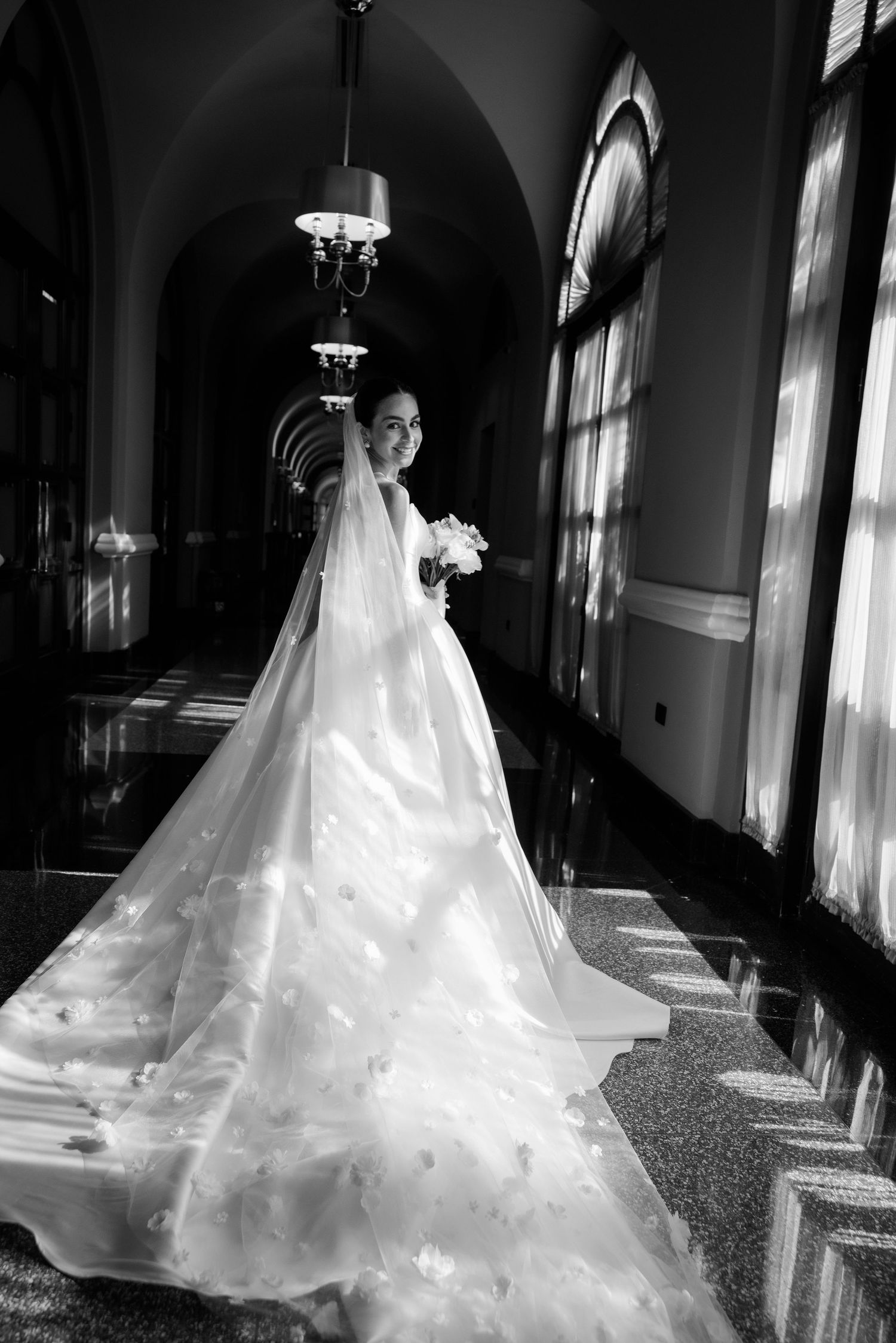 A dramatic black and white photo of a bride in a flowing cathedral-length veil and gown in an elegant hallway.