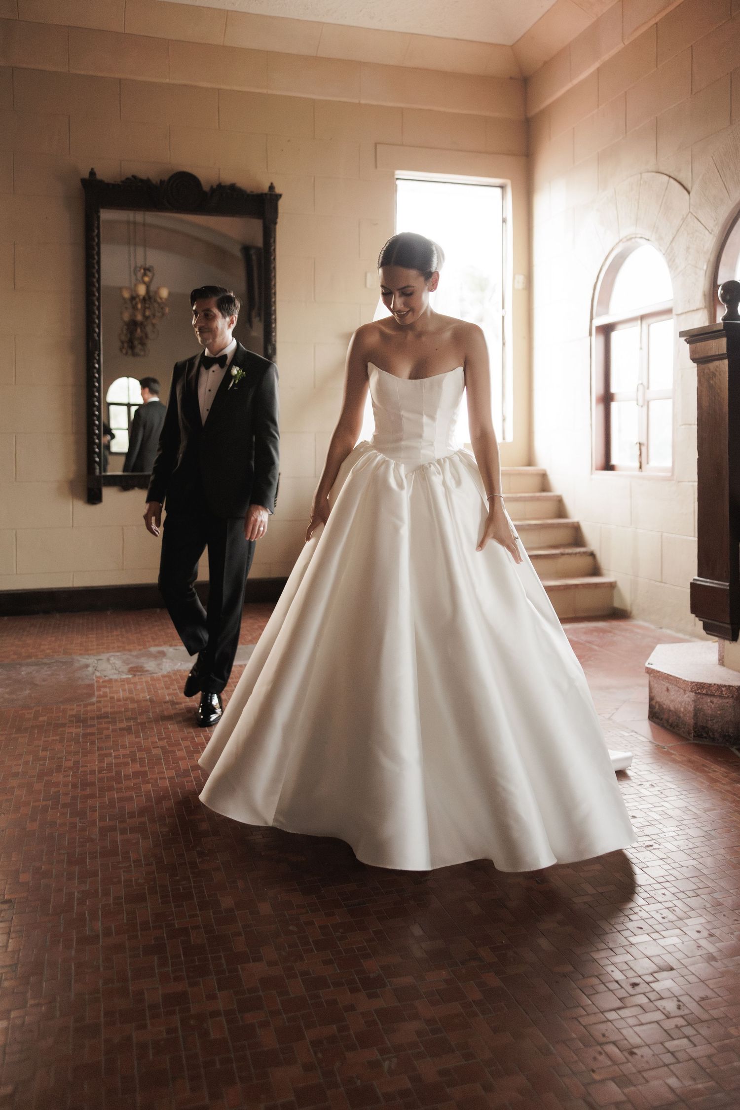 Wedding couple moves through a sunlit indoor space with brick flooring and natural lighting.