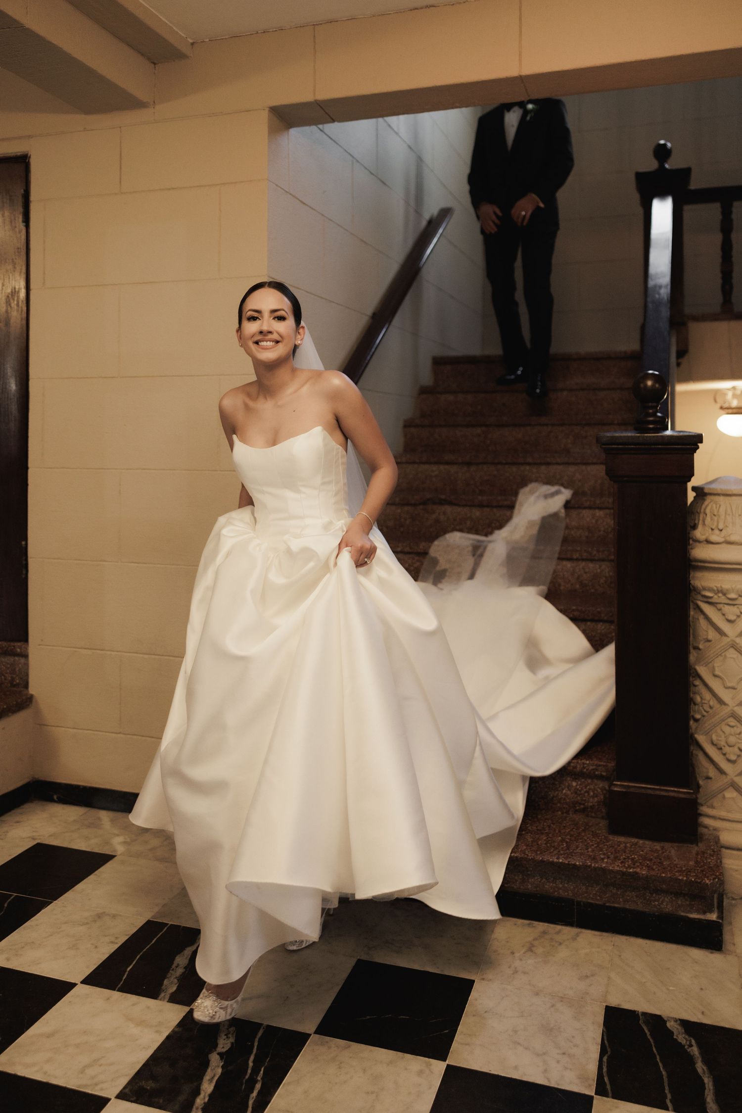 A bride in a white ball gown twirls on a checkered floor near a staircase showing the dress's flowing movement.