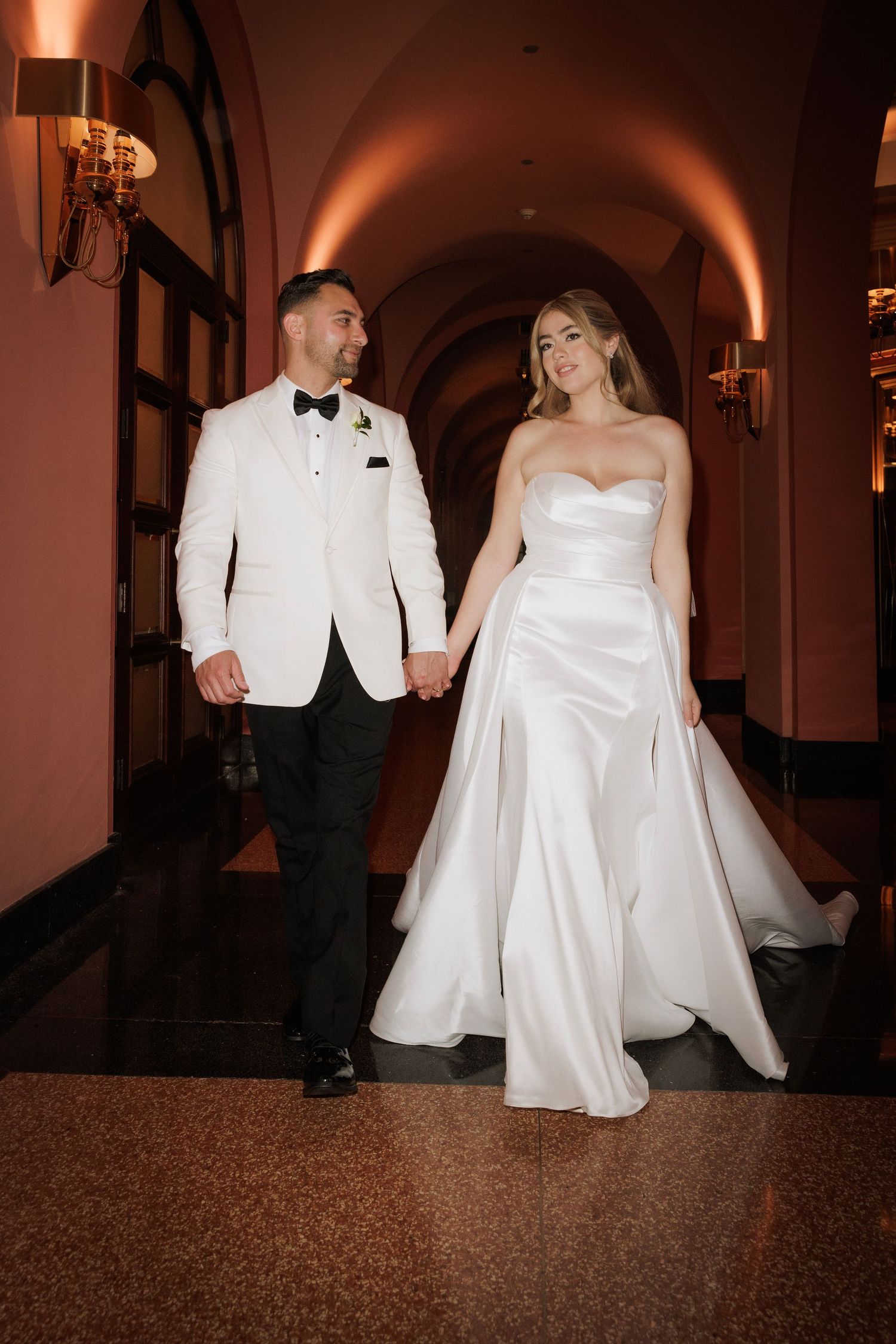 A couple in white formal wear dances in a warmly lit hallway with arched ceilings.