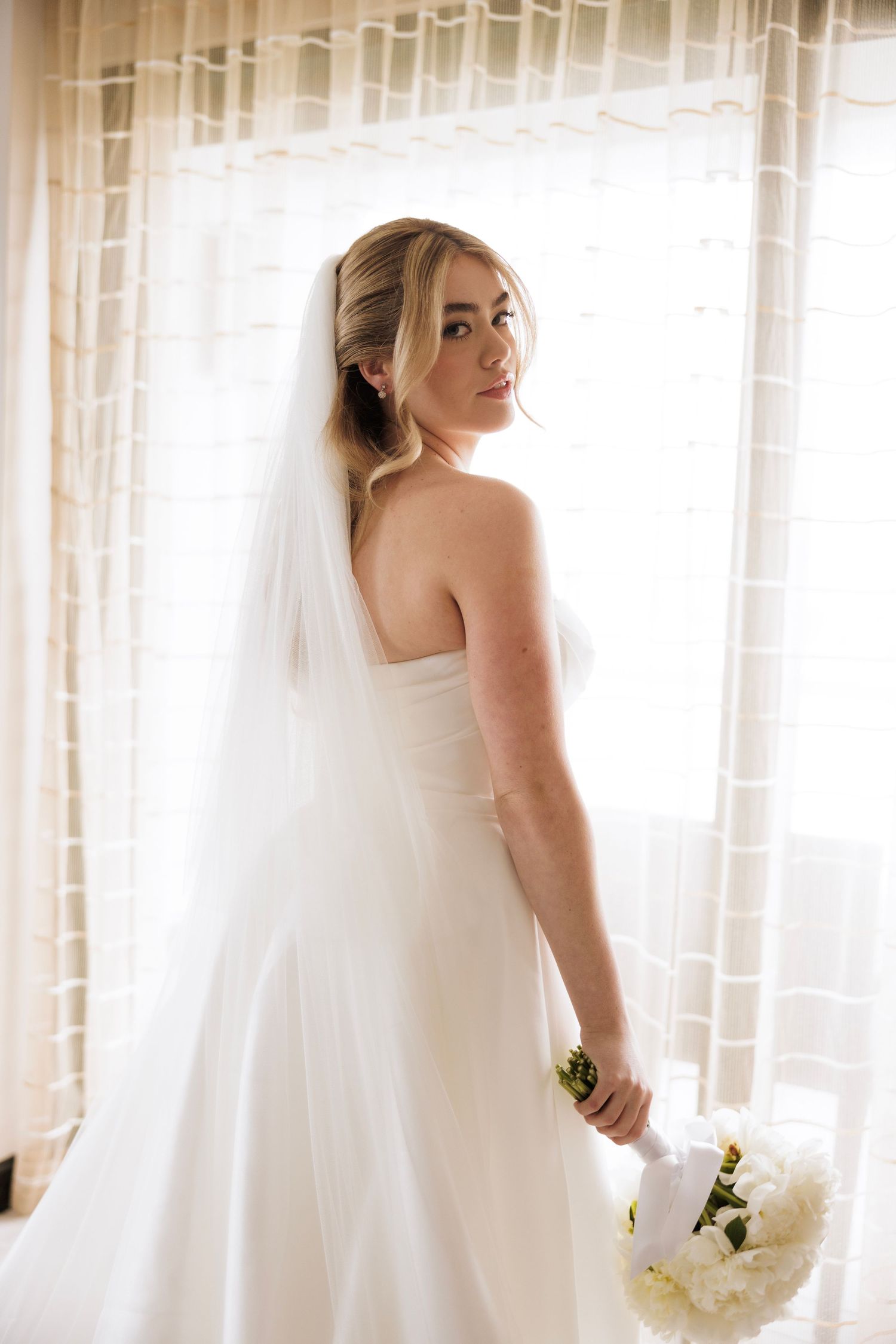 Bride in a flowing white wedding gown poses near sunlit windows holding a white bouquet.