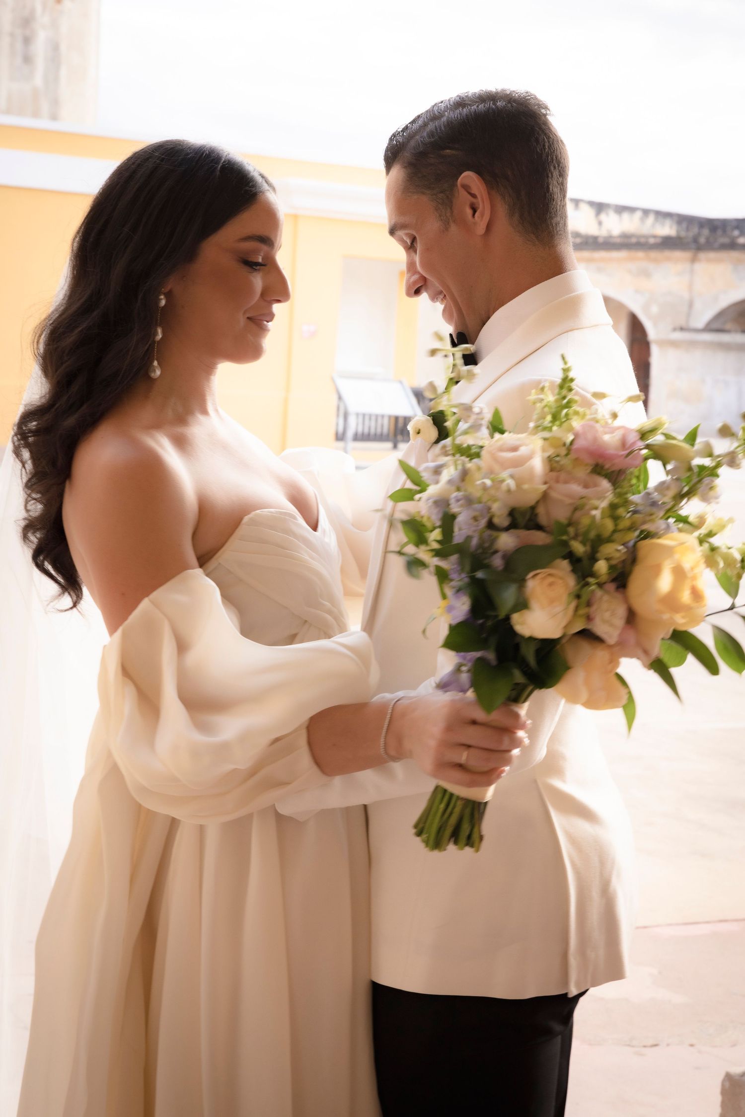 Romantic wedding couple embraces while holding a bouquet of flowers against a light background.
